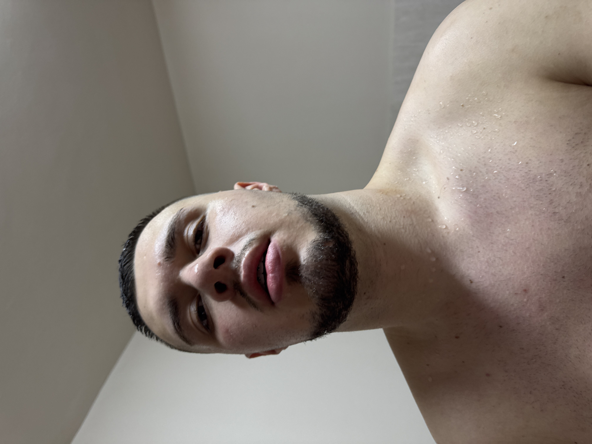 A close-up, low-angle shot shows a man's face and upper body. He has dark hair, a dark beard, and fair skin with water droplets visible on his face and upper chest. The background appears to be a plain, light-colored wall. There is no information in the image to indicate a location in Sofia, Bulgaria, or any other specific location.