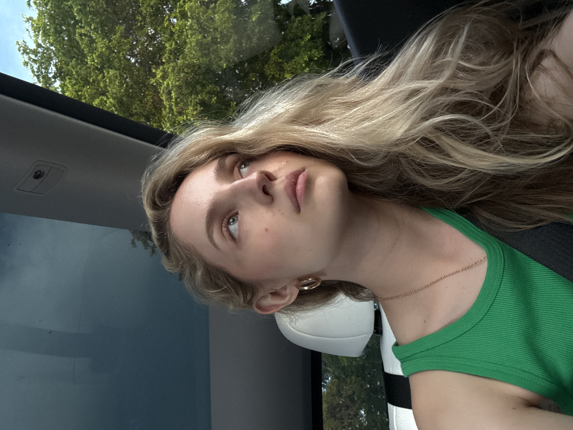 A young woman with blonde, wavy hair is seen from the chest up, looking to her right. She is wearing a green tank top and a gold necklace. She has large hoop earrings and her seatbelt is visible across her chest. The background shows the interior of a car with a sunroof and a view of green trees and a blue sky outside.