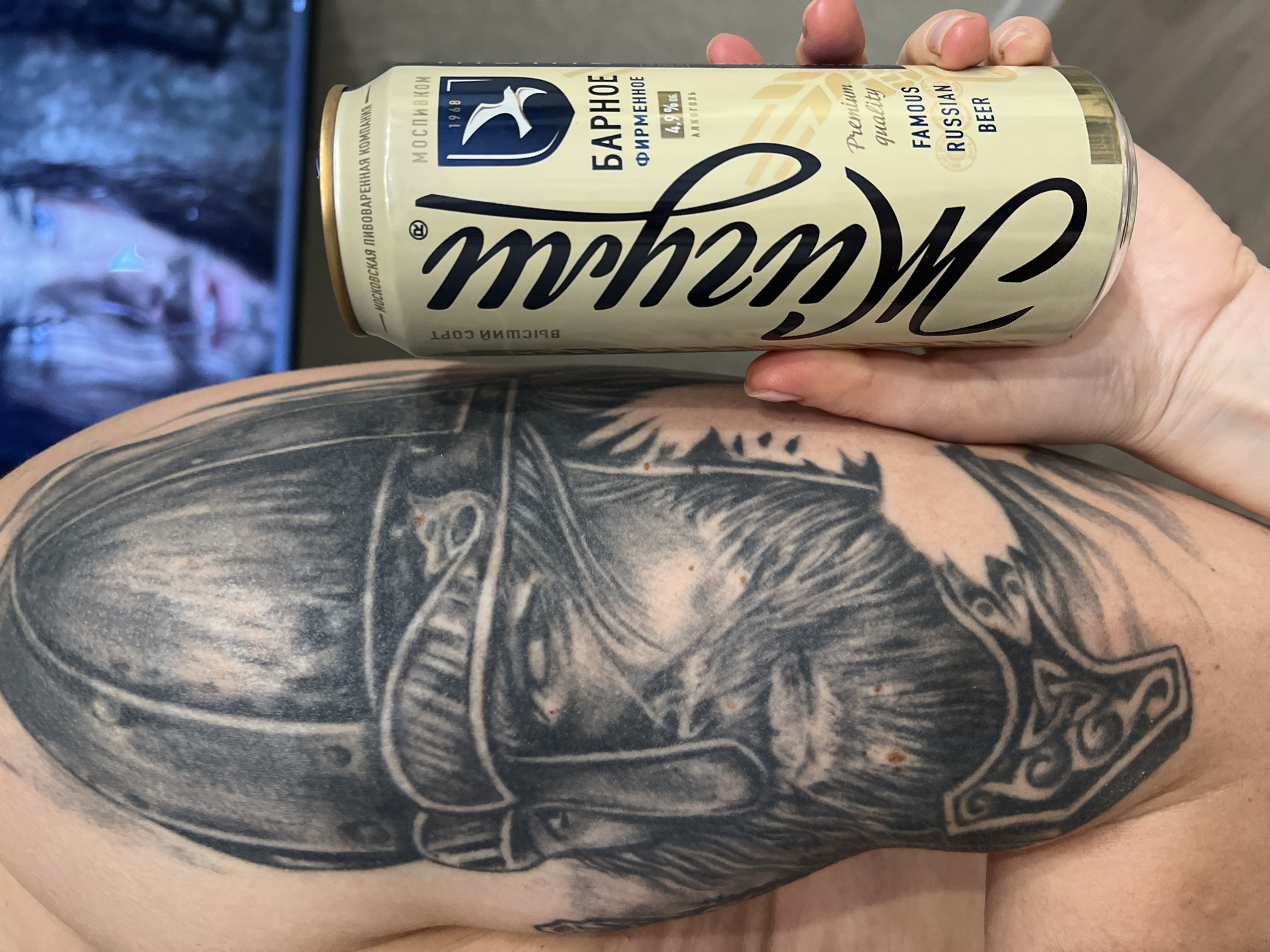 An indoor, close-up shot captures a person's arm holding a light beige beer can against a blurry background that includes a reflective surface, possibly a television screen showing a person's face. The arm features a prominent black and grey tattoo on the bicep/upper arm, depicting a detailed warrior's helmet and, further towards the shoulder, a Mjolnir (Thor's hammer) symbol with Celtic knotwork. The skin shows some freckles or minor blemishes.
The beer can is held by the fingers and thumb of one hand. Its label is primarily light beige with dark blue/black text. Visible Russian text includes "Жигули" (Zhiguli) in a large script font, "БАРНОЕ ФИРМЕННОЕ" (Barnoe Firmennoe), "4,9% об. АЛКОГОЛЬ" (4.9% vol. ALCOHOL), "МОСКОВСКАЯ ПИВОВАРЕННАЯ КОМПАНИЯ" (Moscow Brewing Company), "МОСПИВКОМ" (Mospivkom), and the year "1968". English text reads "Premium quality" and "FAMOUS RUSSIAN BEER". A partially visible, upside-down label also states "ВЫСШИЙ СОРТ" (Vysshiy Sort). The can has a metallic gold-colored top rim. The overall composition highlights both the beverage and the arm tattoo.