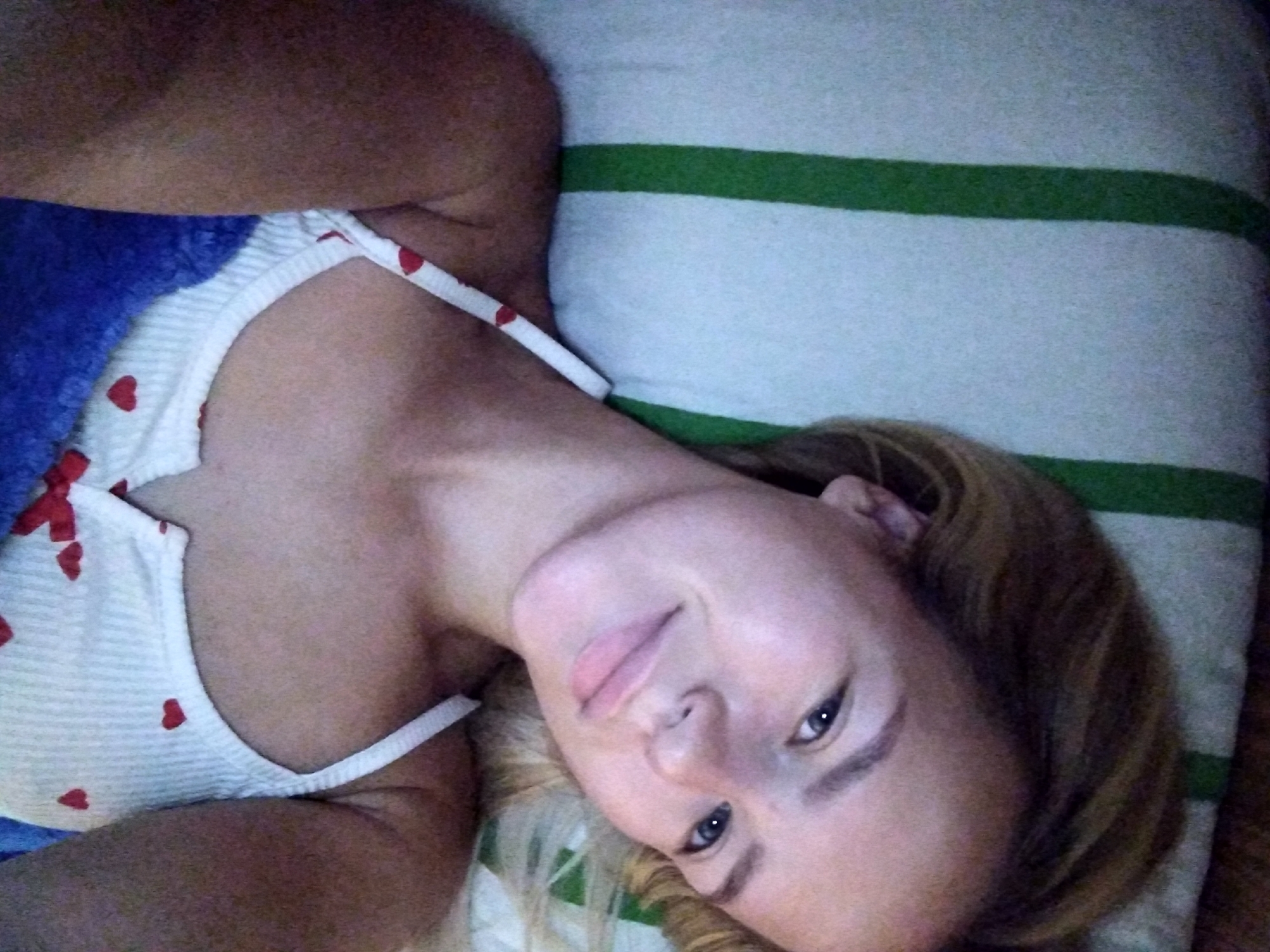 A young woman with blonde hair is lying in bed, looking at the camera. She is wearing a white camisole with red hearts on it. She is under a blue blanket, and there is a striped pillow behind her. The image is taken from a slightly elevated angle.