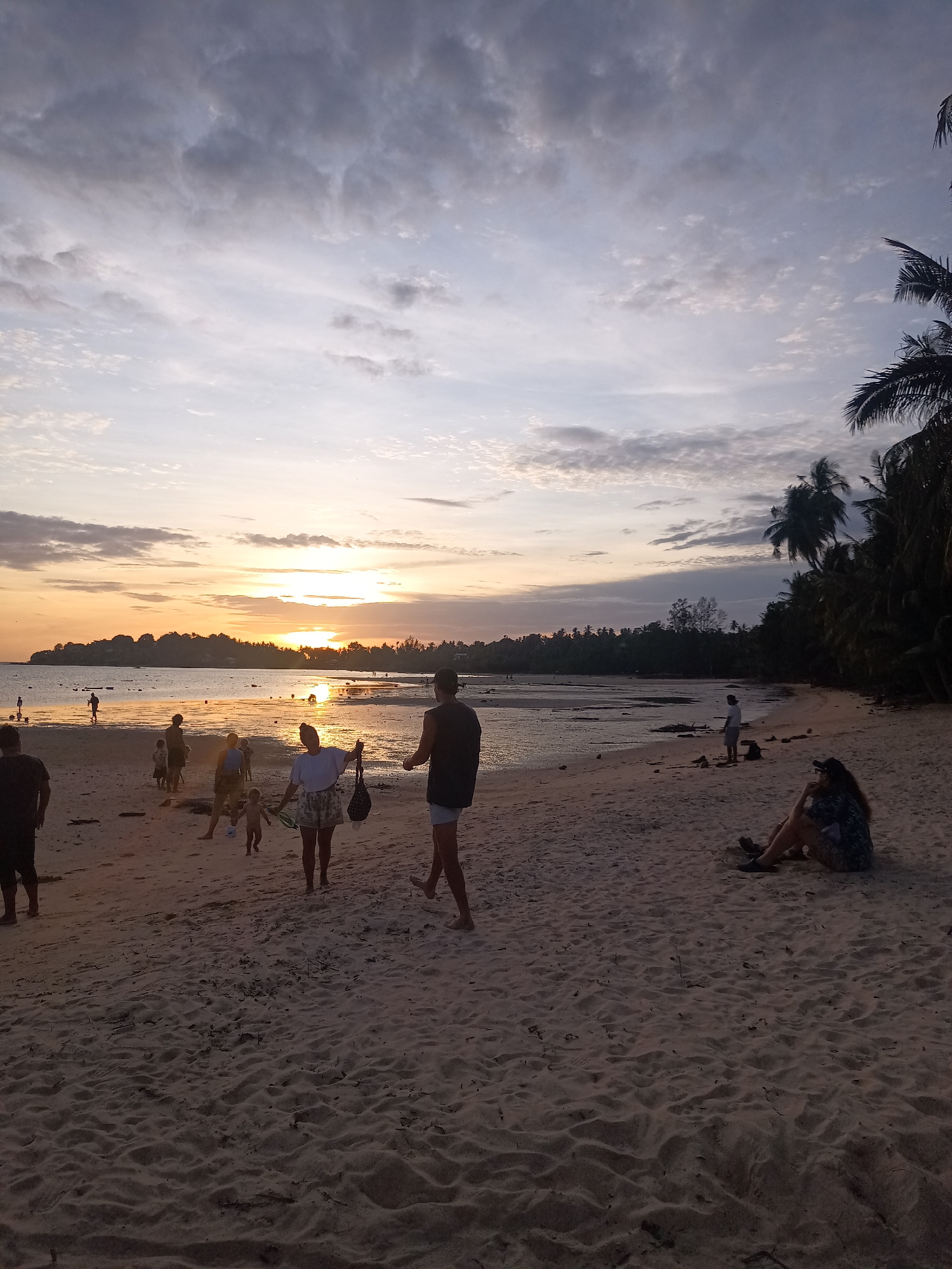 The image depicts a sunset scene on a sandy beach in Koh Phangan, Thailand. Several people are scattered across the sand and near the water's edge. In the foreground, a person is sitting on the beach facing away from the camera. Further up the beach, a group of people are walking and interacting. In the middle ground, more individuals are visible near the water, with some wading in the shallow sea. The background shows a tree-lined horizon with the sun setting over the water, casting a warm glow. Palm trees are visible on the right side of the image, suggesting a tropical location. The sky is filled with clouds, some of which are illuminated by the setting sun.