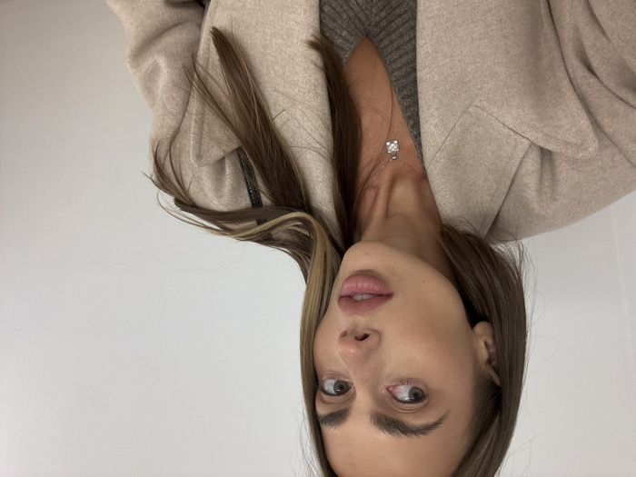 A woman with brown hair and green eyes poses in front of a white wall. She wears a beige coat over a grey sweater, and a silver necklace with a square pendant is visible. Her hair is styled with face-framing highlights, and her lips are a natural pink. The photo is taken from a slightly upward angle, capturing her face and upper body.