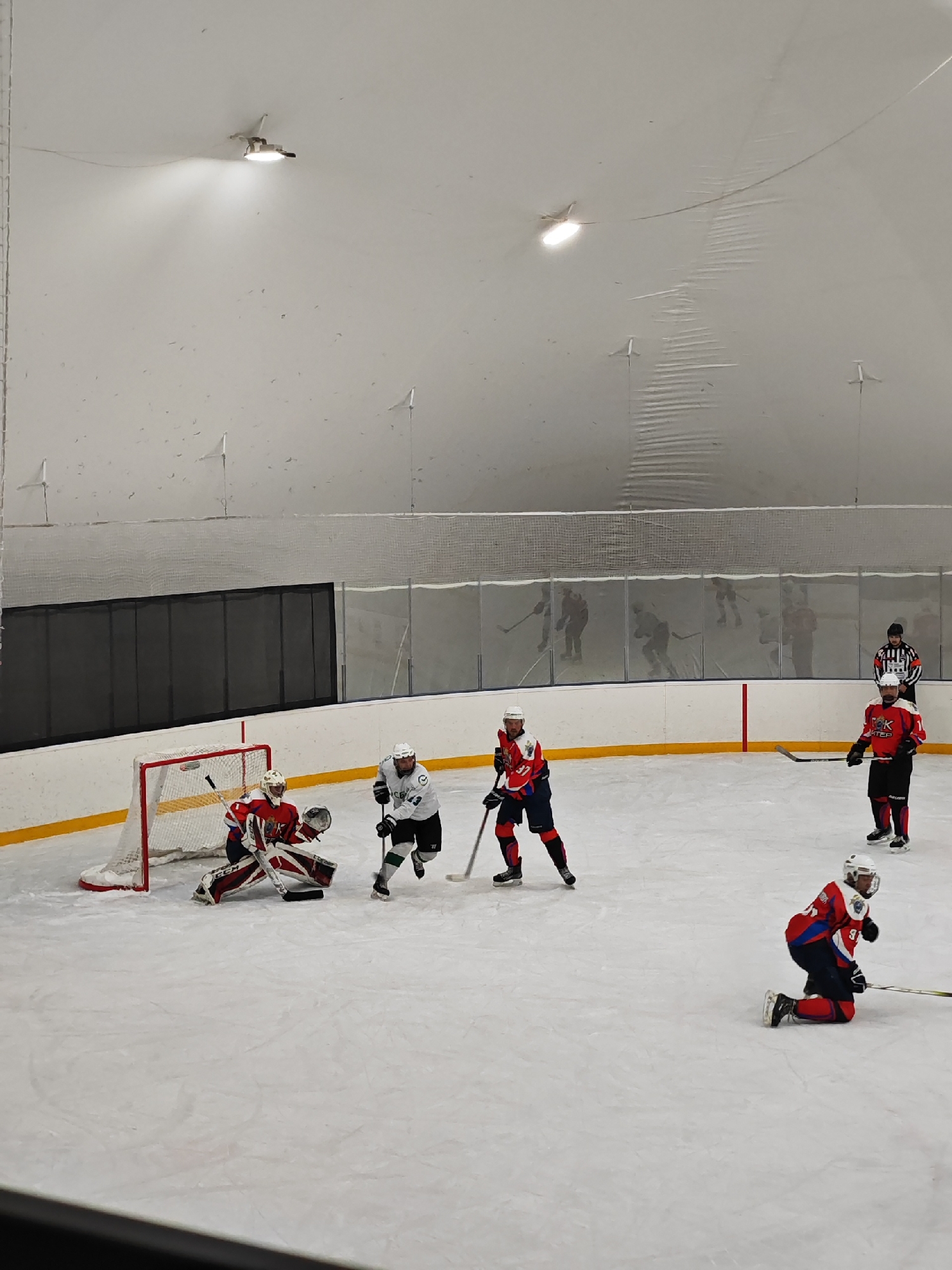 The image shows a hockey game in progress at an indoor ice rink. A goalie in red and white gear defends the net, while an opposing player in white and green approaches with the puck. Two other players in red, white, and blue are on the ice, one standing and one kneeling. In the background, other skaters are visible through the protective netting along with a referee. The location is in Saint Petersburg, Russia. The rink is covered by a white dome-shaped roof and lit by overhead lights.
