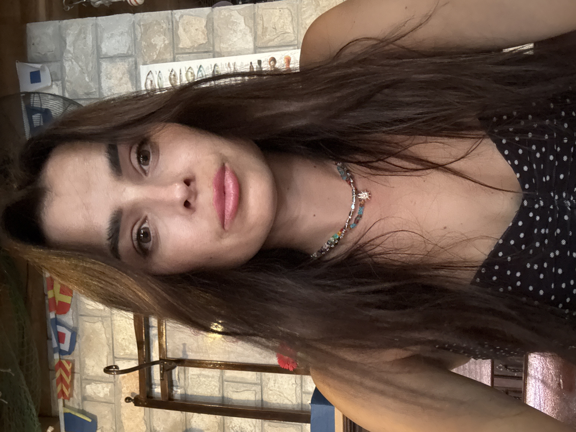 A woman with long, dark brown hair takes a selfie. She is wearing a black and white polka dot top and a multi-colored beaded necklace with a sun pendant. The background appears to be an outdoor or semi-outdoor setting with a brick wall, nautical flags, and a rack.