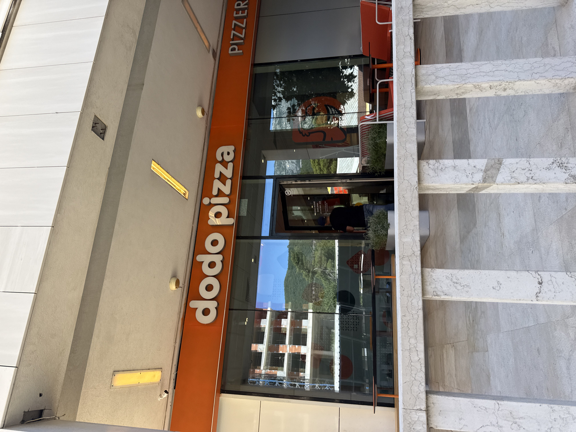 The image shows the exterior of a Dodo Pizza restaurant in Budva, Montenegro, as indicated by the signage and the architectural style. The restaurant has a large glass storefront with orange trim. The name "dodo pizza" is prominently displayed in white lettering on the orange trim. To the right of the Dodo Pizza sign, a sign for "PIZZERIA" is visible.
Inside the restaurant, through the glass, a construction site is reflected, showing an unfinished building with exposed brick and scaffolding. There are also tables and chairs visible, both inside and outside the restaurant. Outside, a man in a black t-shirt and jeans is standing near the entrance, possibly an employee or a customer. Orange benches and planters with greenery are also present on the outdoor seating area. The overall environment suggests a sunny day with clear skies, with a backdrop of hills and trees visible through the glass. The foreground is dominated by a modern-looking white marble railing and paved ground.