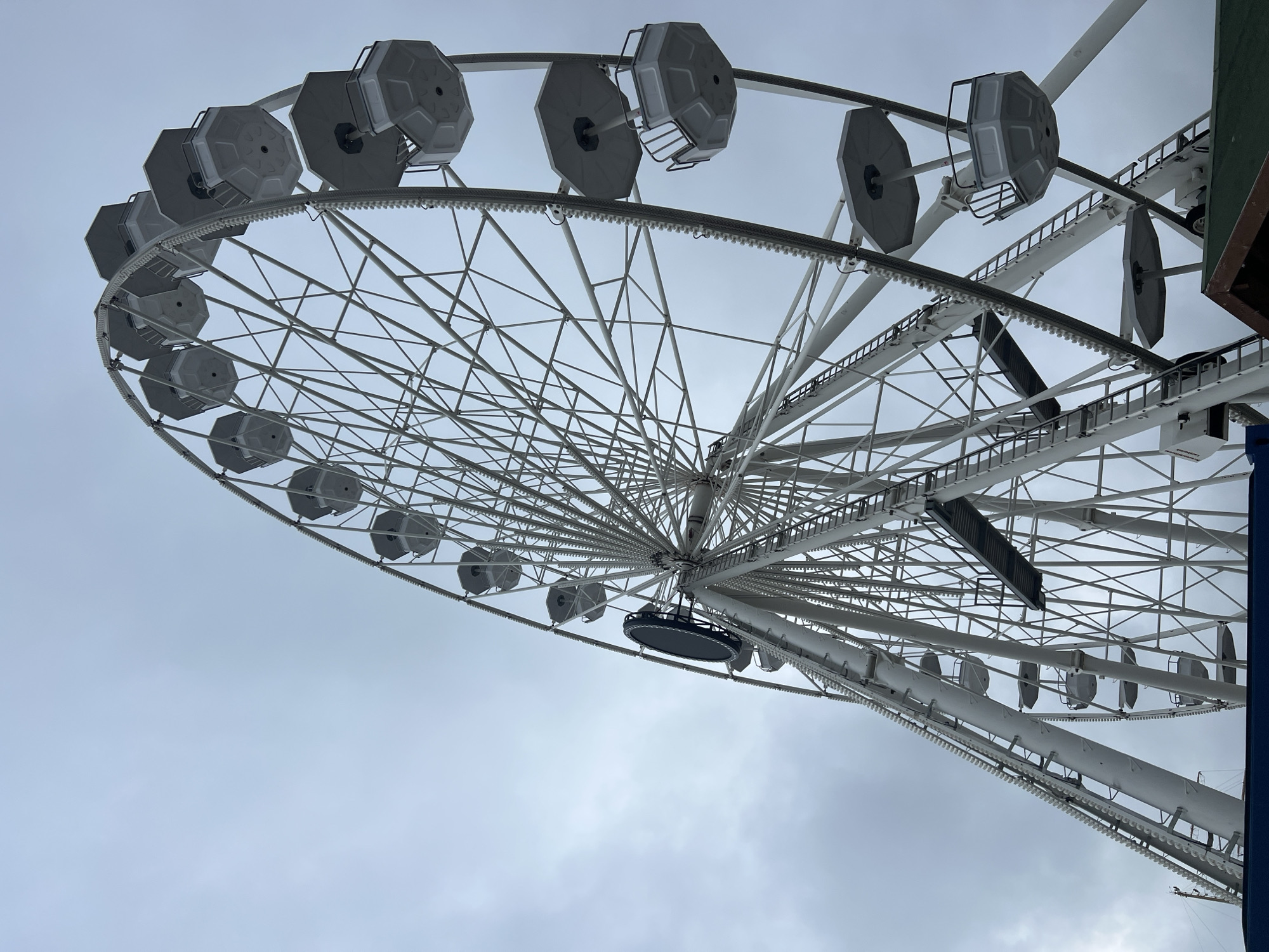 The image presents a low-angle view looking up at a large Ferris wheel against an overcast sky. The Ferris wheel, predominantly white, fills the majority of the frame, showcasing its intricate network of spokes radiating from a central hub. Numerous hexagonal-shaped cabins, or gondolas, are suspended along the outer circumference of the wheel. These cabins are gray and white, some appearing to be open at the top or front.
The sky is a uniform light gray, indicating a cloudy or overcast day, with no direct sunlight visible. The bright white structure of the Ferris wheel stands out against the muted sky. On the far right side of the image, a dark green and brown structure, possibly part of a building or support mechanism, is partially visible. The Ferris wheel appears stationary. No people are visible within the cabins or around the structure, nor is any visible text present on the wheel or surrounding elements. The scene captures the imposing scale and mechanical design of the attraction, located in Varna, Bulgaria.