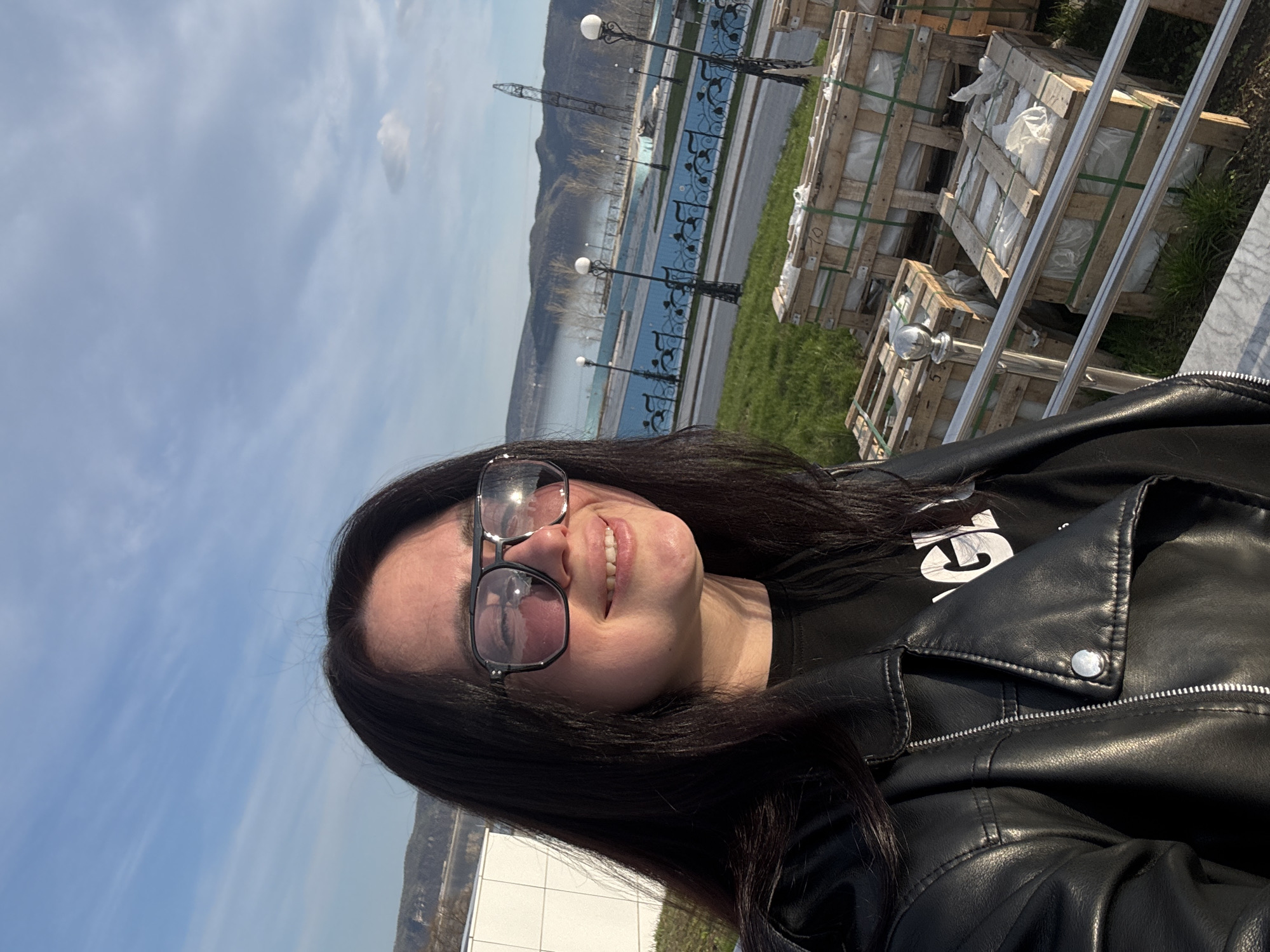 The image captures a selfie of a young woman taken outdoors on a bright, sunny day. The woman, positioned prominently in the foreground, is smiling widely and looking directly at the camera. She has long, dark hair and is wearing dark-rimmed sunglasses, a black leather jacket with a silver snap button and zipper details, and a black t-shirt. The sunglasses reflect the clear sky and surroundings.
Behind her, a scenic waterfront landscape unfolds. A calm body of water, possibly a river or a canal, is visible across the middle ground. Along the water's edge, there is a promenade or paved area adorned with multiple black decorative lampposts and an ornate black metal railing featuring blue patterned elements. Beyond the waterfront, dark mountains or hills rise under a light blue sky dotted with scattered white clouds. A tall, slender structure, resembling a crane or communication tower, stands in the distance near the mountains.
To the right of the woman, several wooden crates, some wrapped in white plastic sheeting, are stacked on a patch of green grass. These appear to be materials or goods, possibly for construction or delivery. Metallic fixtures, possibly part of another railing, are partially visible in the bottom right corner.
The time of day is daytime, indicated by the bright sunlight. The weather is clear and pleasant. A large white letter "G" is visible on the woman's black t-shirt, part of a larger, mostly obscured word. On one of the wooden crates, the number "10" is discernible, and another appears to have "5" or "5B" marked on it. The overall mood conveyed is cheerful and relaxed.