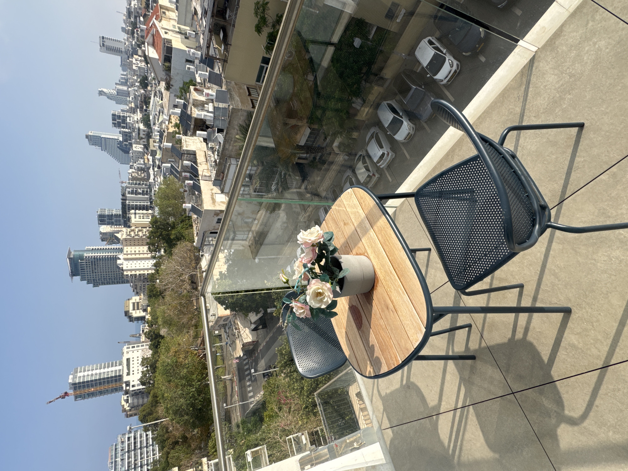 A high-angle shot showcases a round wooden table with a potted arrangement of light-colored roses, alongside a metal chair. The location is likely a balcony or terrace, judging from the glass barrier visible. Beyond the barrier, the skyline of Tel-Aviv, Israel is visible, including buildings, trees, and distant construction. The overall lighting suggests a sunny day.