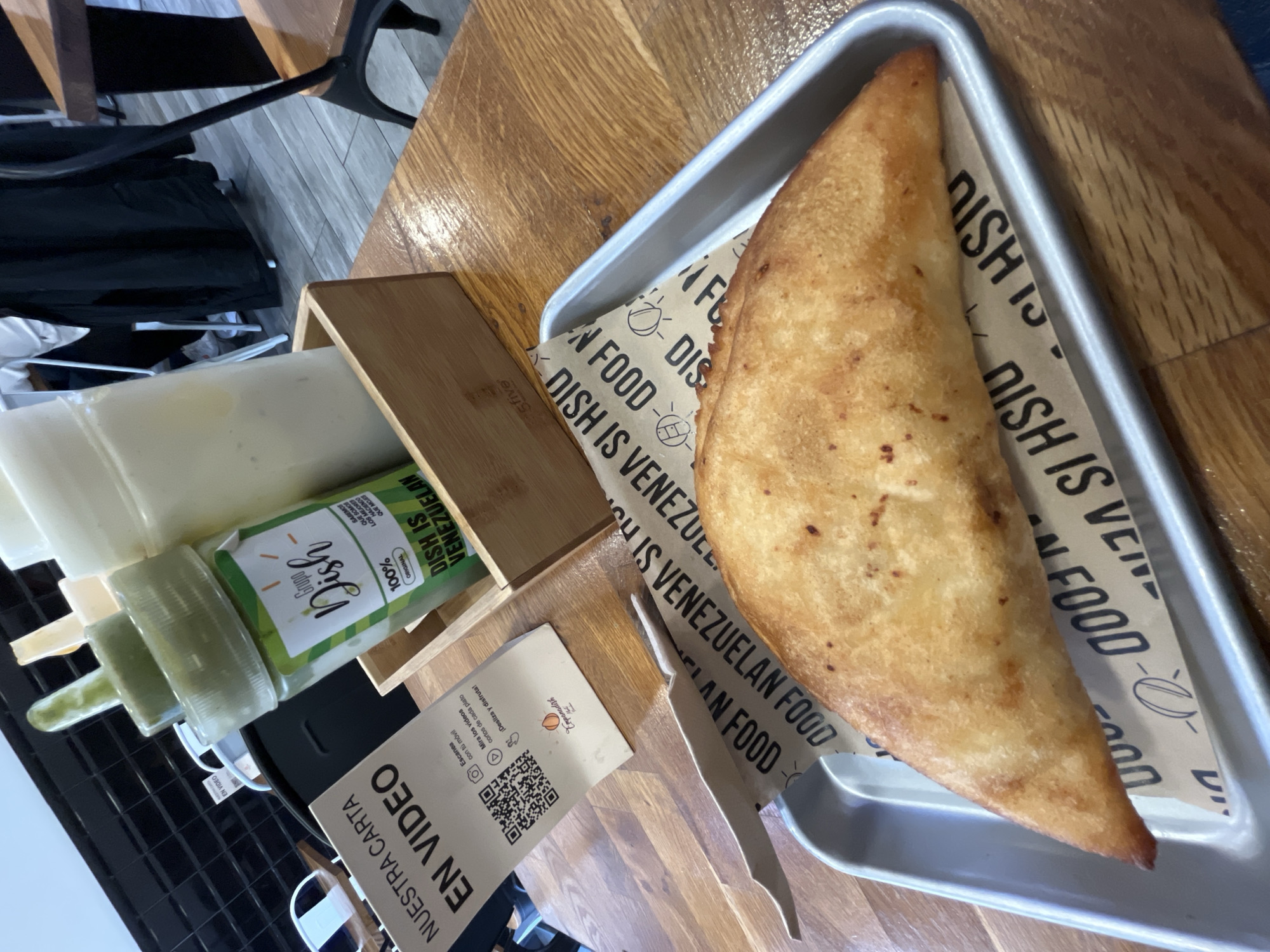 The image shows a close-up of a metal serving tray holding a large, golden-brown empanada. The empanada is resting on a paper liner with the words "DISH IS VENEZUELAN FOOD" printed repeatedly. To the left of the tray, there is a wooden holder containing several squeeze bottles, one of which is green. Next to the wooden holder is a business card with a QR code and the words "NUESTRA CARTA EN VIDEO." The background consists of a wooden table, a chair, and a black grid-like surface. The name "Empanadish" on the business card and the words "DISH IS VENEZUELAN FOOD" suggest that the food is Venezuelan cuisine. This is the restaurant Empanadish, which has two locations in Barcelona, Spain.
