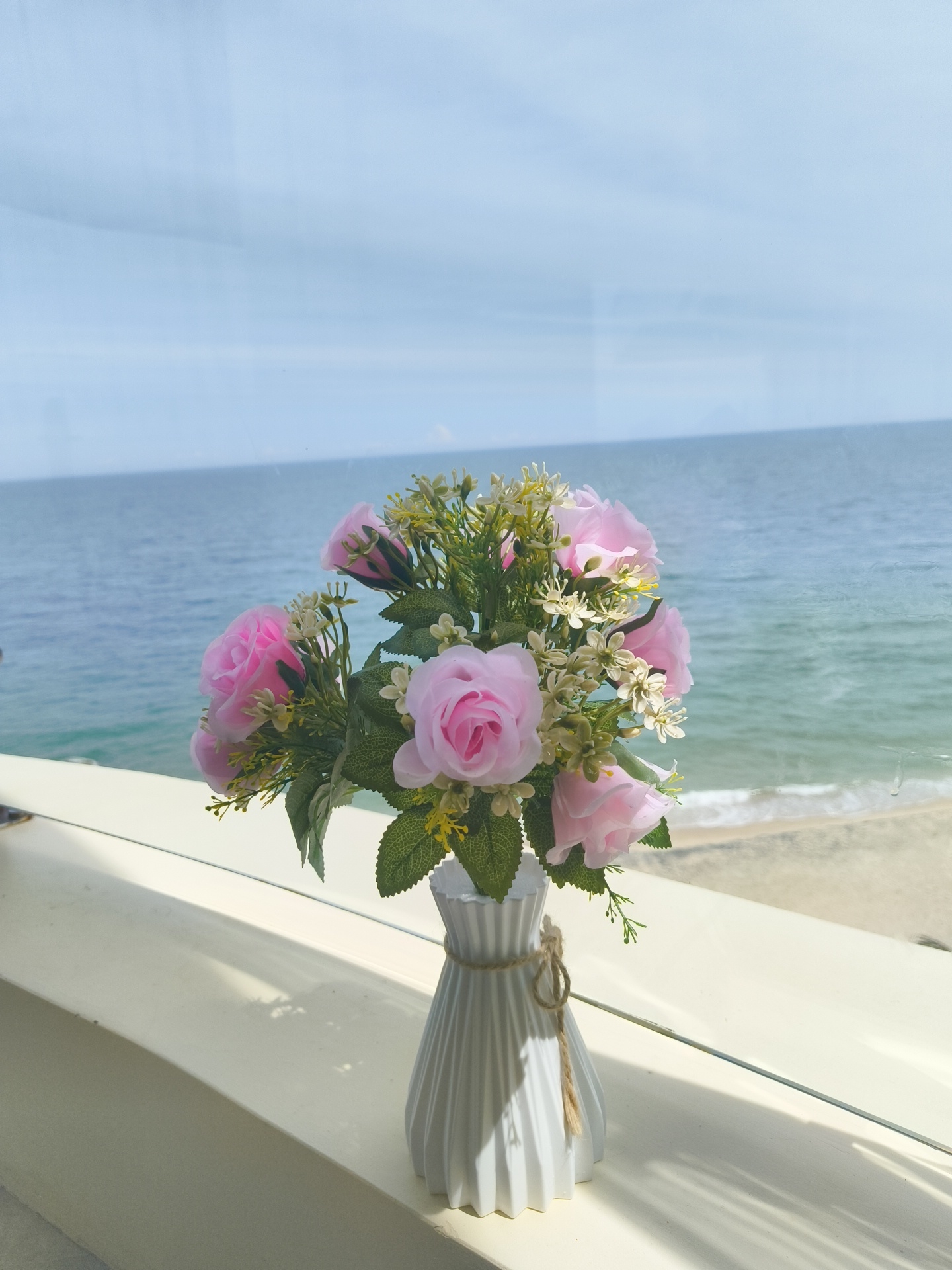 A bouquet of artificial pink roses with green foliage and tiny white flowers is displayed in a white fluted vase tied with jute twine. The vase sits on a cream-colored ledge, which appears to be a windowsill or balcony railing. The ledge casts a shadow from sunlight. In the background, a calm blue ocean meets a sandy beach under a clear, pale blue sky. The scene is likely situated in Phu Thuy Ward, Vietnam, given the context provided, and evokes a serene coastal atmosphere. There are no people or animals present in the image.