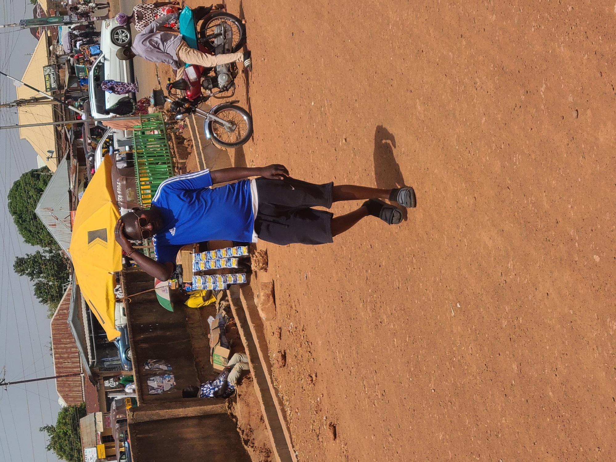 The scene is a street in Jos, Nigeria. The ground is red dirt. A man wearing a blue shirt, black shorts, and sandals stands beside a low wall, possibly a vendor at a roadside stall, shielding his eyes. A yellow umbrella is open near him, casting a shadow. Stacks of small packets are visible on the wall next to him. A motorcycle and a person riding on it are in the background, along with other vehicles and buildings typical of a busy street. Power lines stretch across the sky.