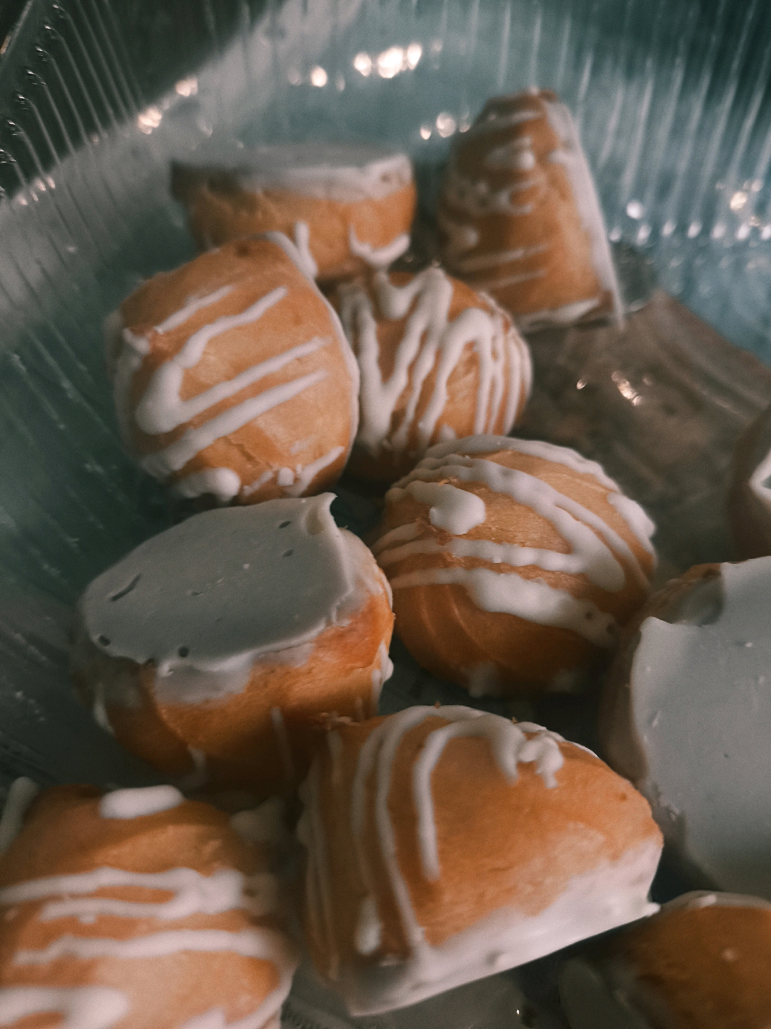 In the city of Слабодскі сельскі Савет, Belarus, a close-up shot captures a clear plastic container filled with an assortment of baked goods. The items appear to be small, round pastries, some of which are partially covered in white icing, while others are drizzled with thin white lines. The pastries exhibit an orange hue, suggesting they might be flavored or colored. The background is blurred, but hints of light reflections on the plastic container and a subtle teal color suggest an indoor environment, possibly a kitchen or a display case. The lighting is dim, creating a slightly moody atmosphere.