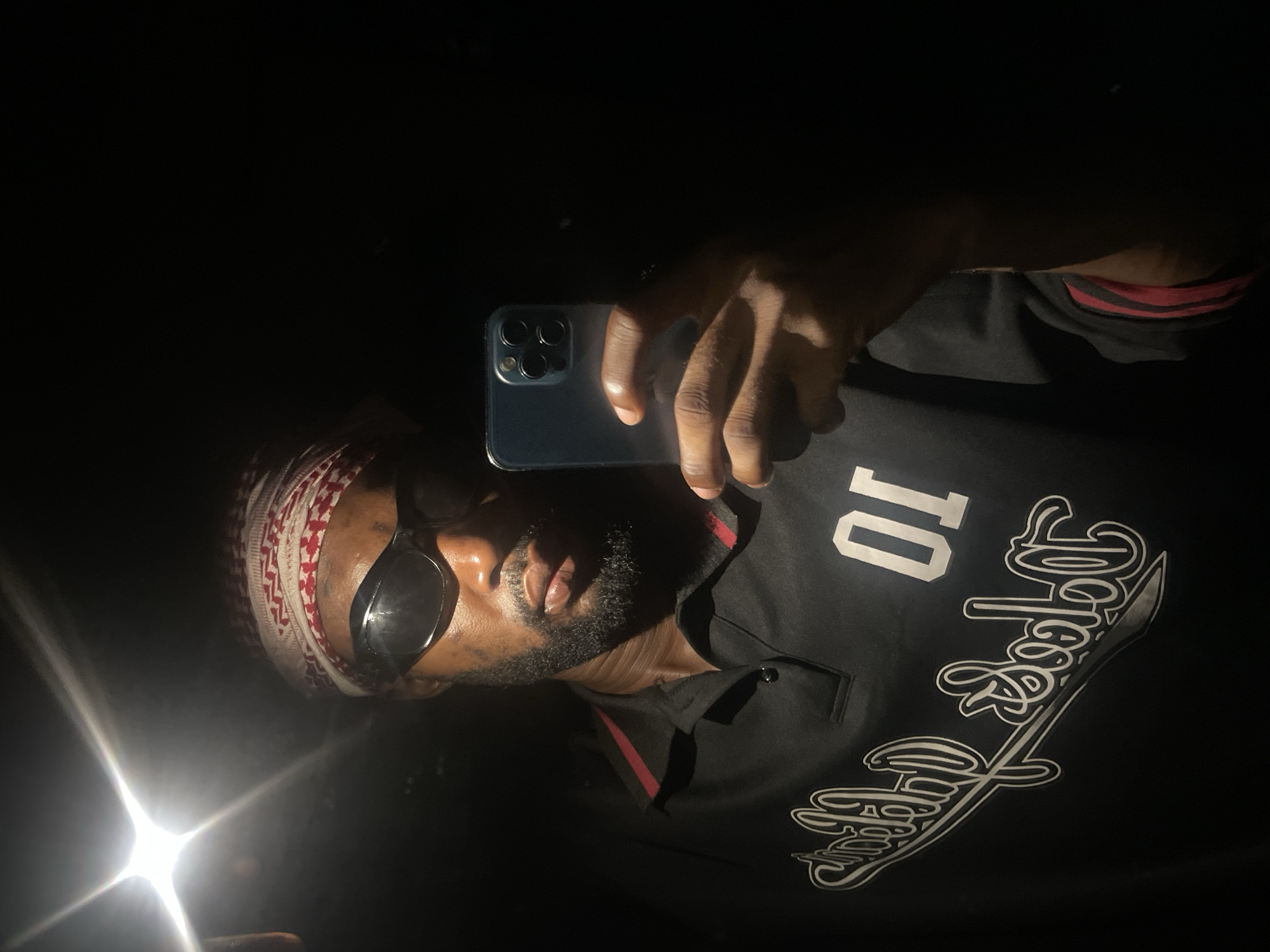A man is holding a smartphone up to take a picture of himself. He is wearing a red and white patterned head covering, sunglasses, and a black shirt with white lettering and the number "10" on it. He has a beard. The environment is dark, and there is a bright light source coming from the lower left of the frame. The location, Lokoja, Nigeria, cannot be confirmed as there are no visible landmarks.