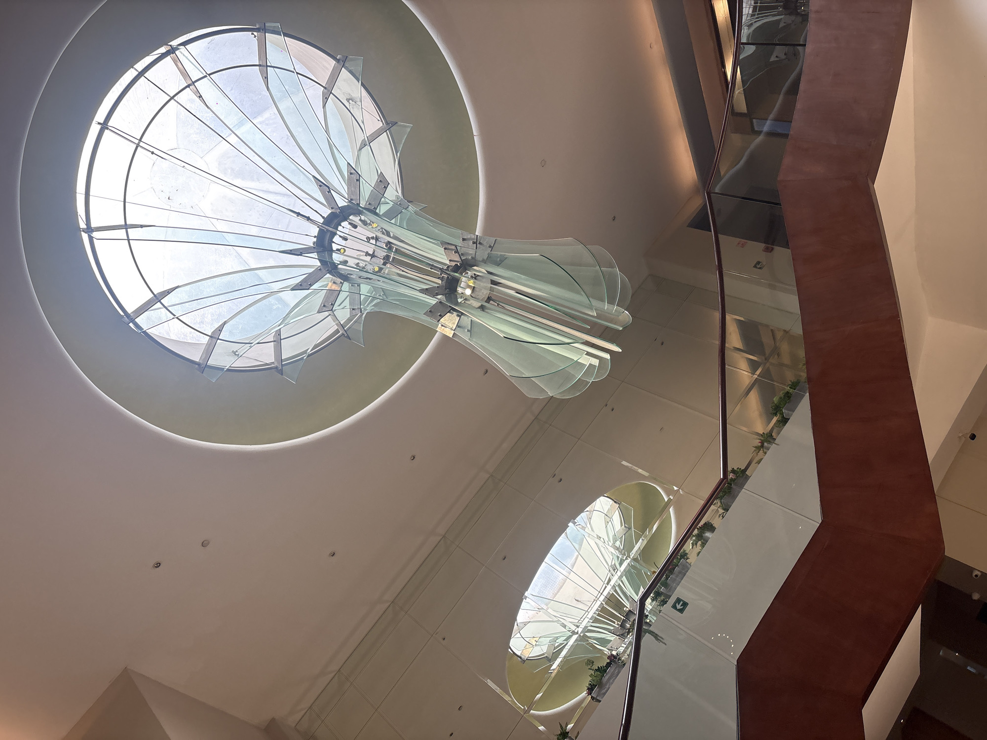 The image shows an interior view of a building in Mexico City, Mexico, featuring a high ceiling with two circular skylights. A decorative glass chandelier hangs beneath the upper skylight. To the right, a multi-story structure is visible, possibly an atrium or elevator shaft, with wood and glass panels. The overall color palette is light, with predominantly white and light beige surfaces, contrasted by the brown wood of the structure.