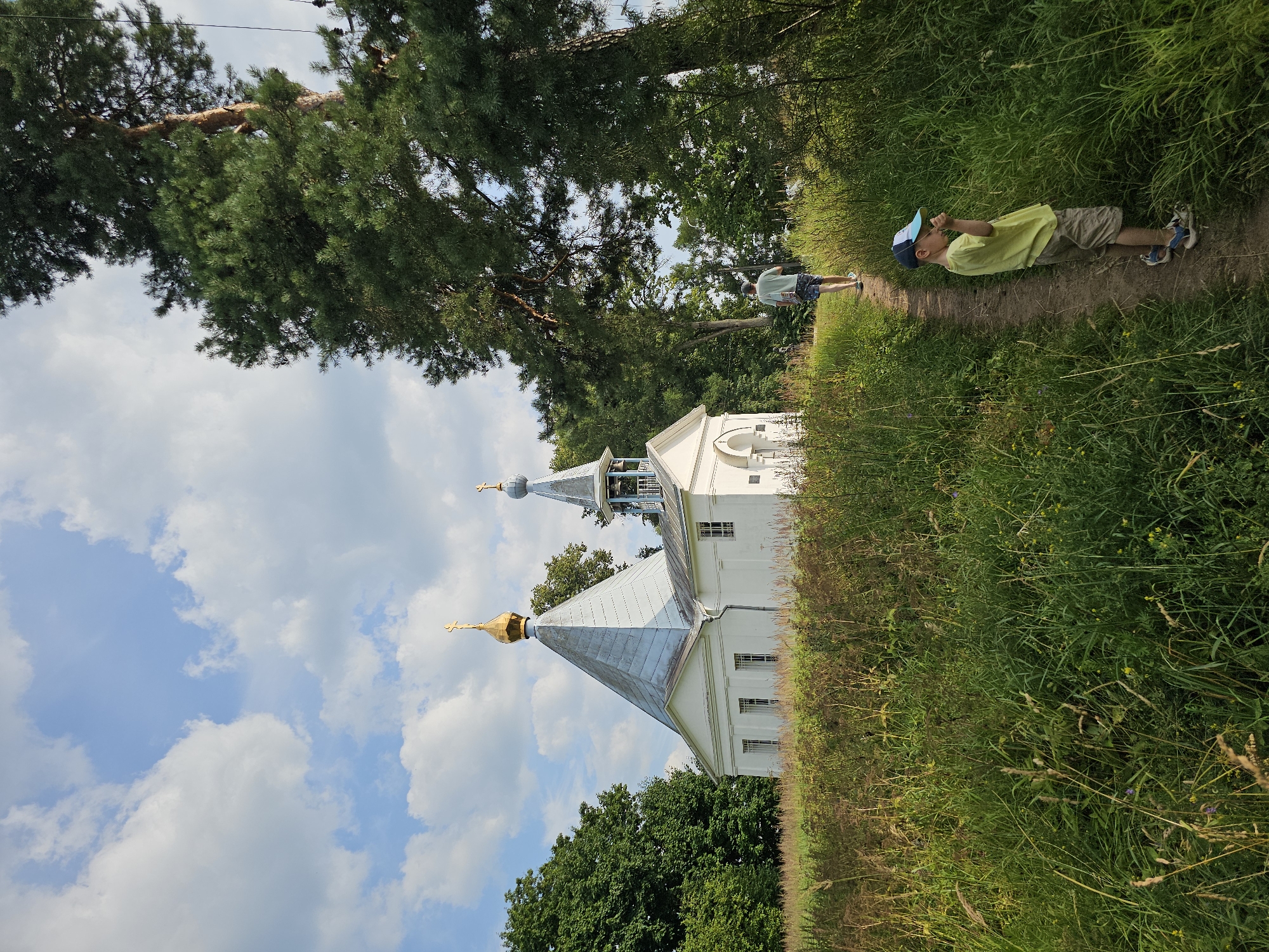 In the village of Ulitkino, Russia, a white Orthodox church with a golden dome and a bell tower stands on a grassy hill. A dirt path leads away from the church, bordered by tall grass and trees. A young boy in a yellow shirt and a blue cap walks down the path, facing away from the camera. Further up the path, a man in a green t-shirt and shorts walks towards the church. The sky is blue with scattered white clouds.
