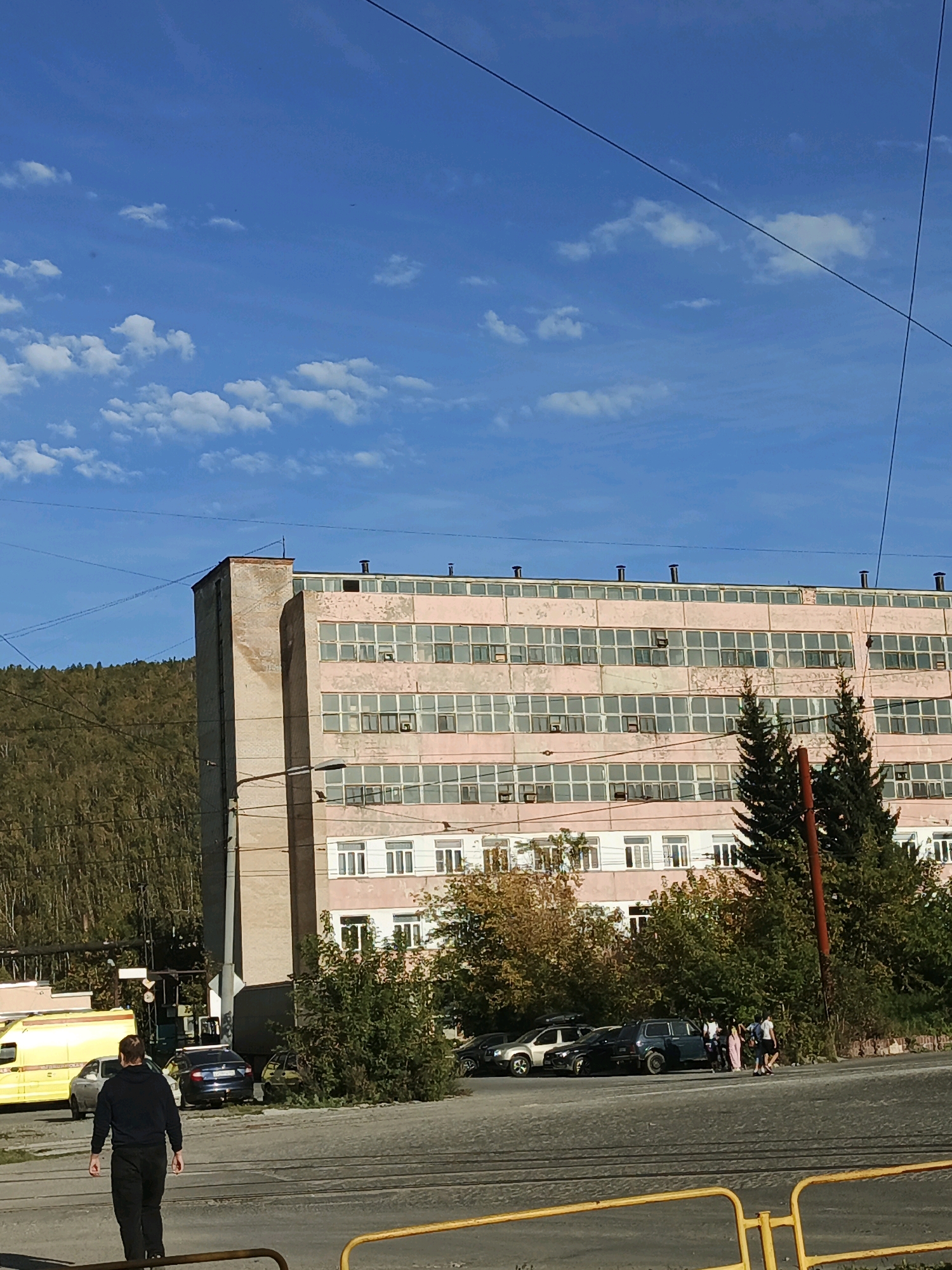 The image depicts an urban scene in Zlatoust, Russia, on a sunny day with a blue sky and scattered white clouds. A large, multi-story building with a light pink facade and numerous windows dominates the central part of the frame. The building appears to be an industrial or administrative facility. In the foreground, a man in a dark hoodie and pants is walking away from the camera across a street with tram tracks. Several cars are parked to the right of the building, and a group of people are standing near them. To the left of the building, a yellow vehicle, possibly an ambulance or a service vehicle, is visible. A forested hillside forms the backdrop to the right of the building. Overhead power lines are visible crisscrossing the sky.