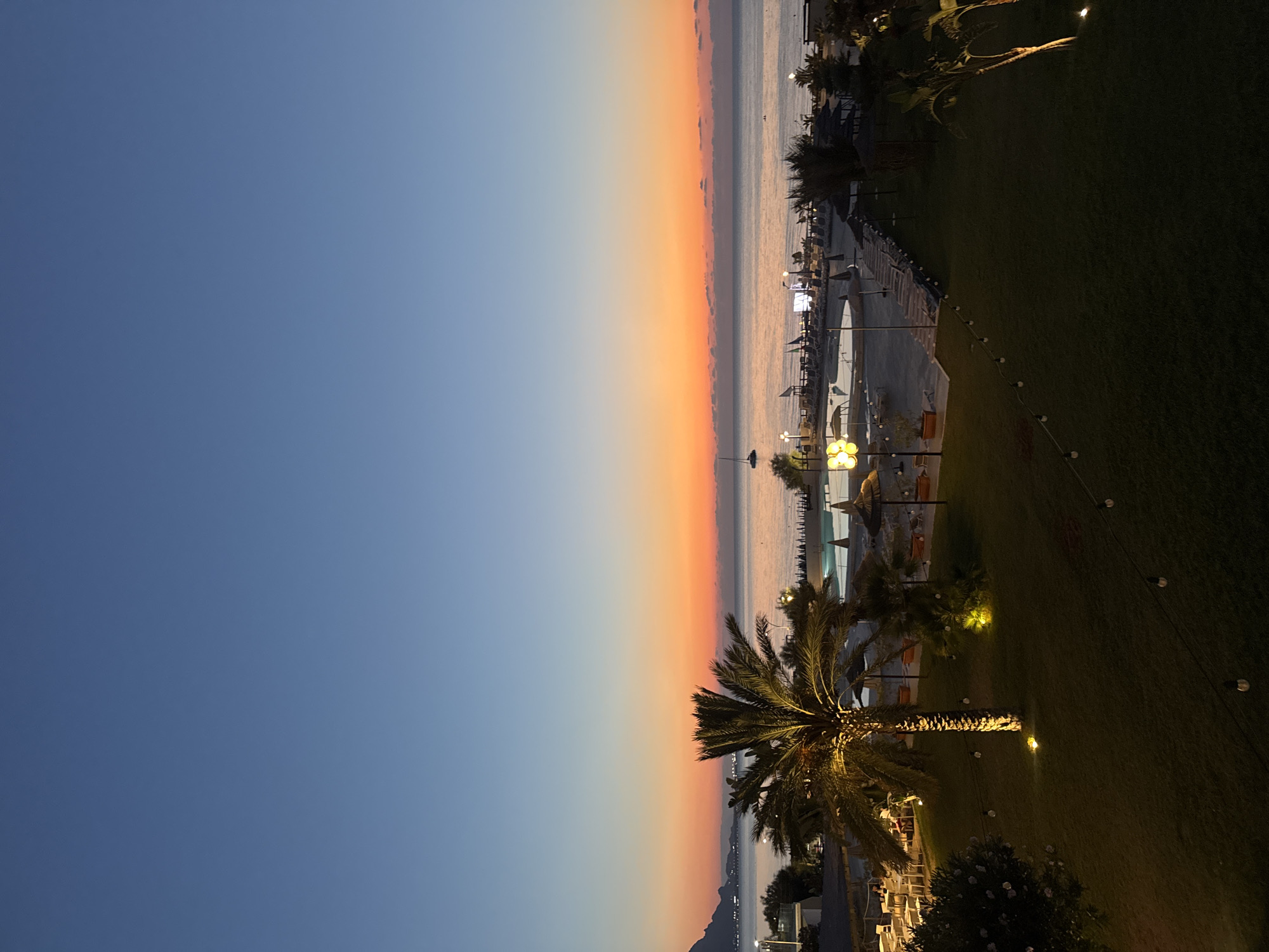 The image captures a serene twilight scene at a beach resort located in Isola delle Femmine, Italy. The sky transitions from a deep blue at the top to a vibrant orange and pink gradient near the horizon, indicating either sunrise or sunset. The calm sea stretches out, with a faint silhouette of mountains visible in the distance.
In the foreground, a well-maintained lawn is illuminated by strategically placed outdoor lights and a string of bulbs. A tall palm tree, its fronds dramatically lit from below, stands prominently in the center-left of the frame. To the left of the palm tree, outdoor dining furniture is set up under a canopy, suggesting a restaurant or lounge area.
Further back, a swimming pool with loungers and umbrellas is visible, hinting at a resort's amenities. Beach chairs and umbrellas are also arranged on the sandy area beyond the pool. A small sailboat is anchored on the water, adding a touch of tranquility to the seascape. The overall atmosphere is peaceful and inviting, characteristic of a coastal resort at dusk.