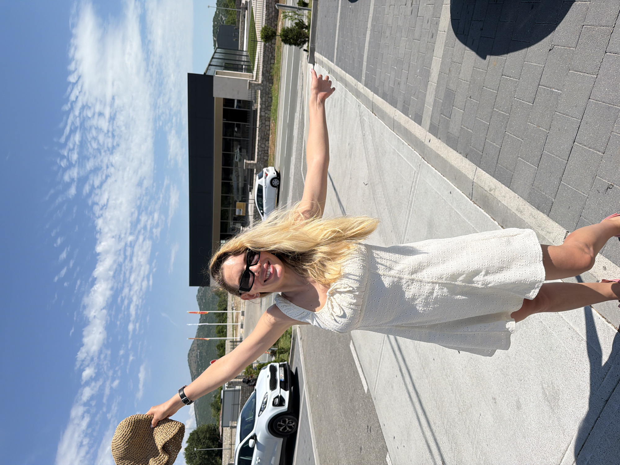 In Bigor, Montenegro, a blonde woman wearing sunglasses and a white eyelet dress stands on a paved sidewalk. She holds a straw hat in her left hand, raised above her head, and extends her right arm to the right, palm open, as if gesturing. Behind her, a white car is parked on the left, and a modern building with dark glass windows stands in the background, flanked by trees and hills under a bright blue sky with scattered white clouds.