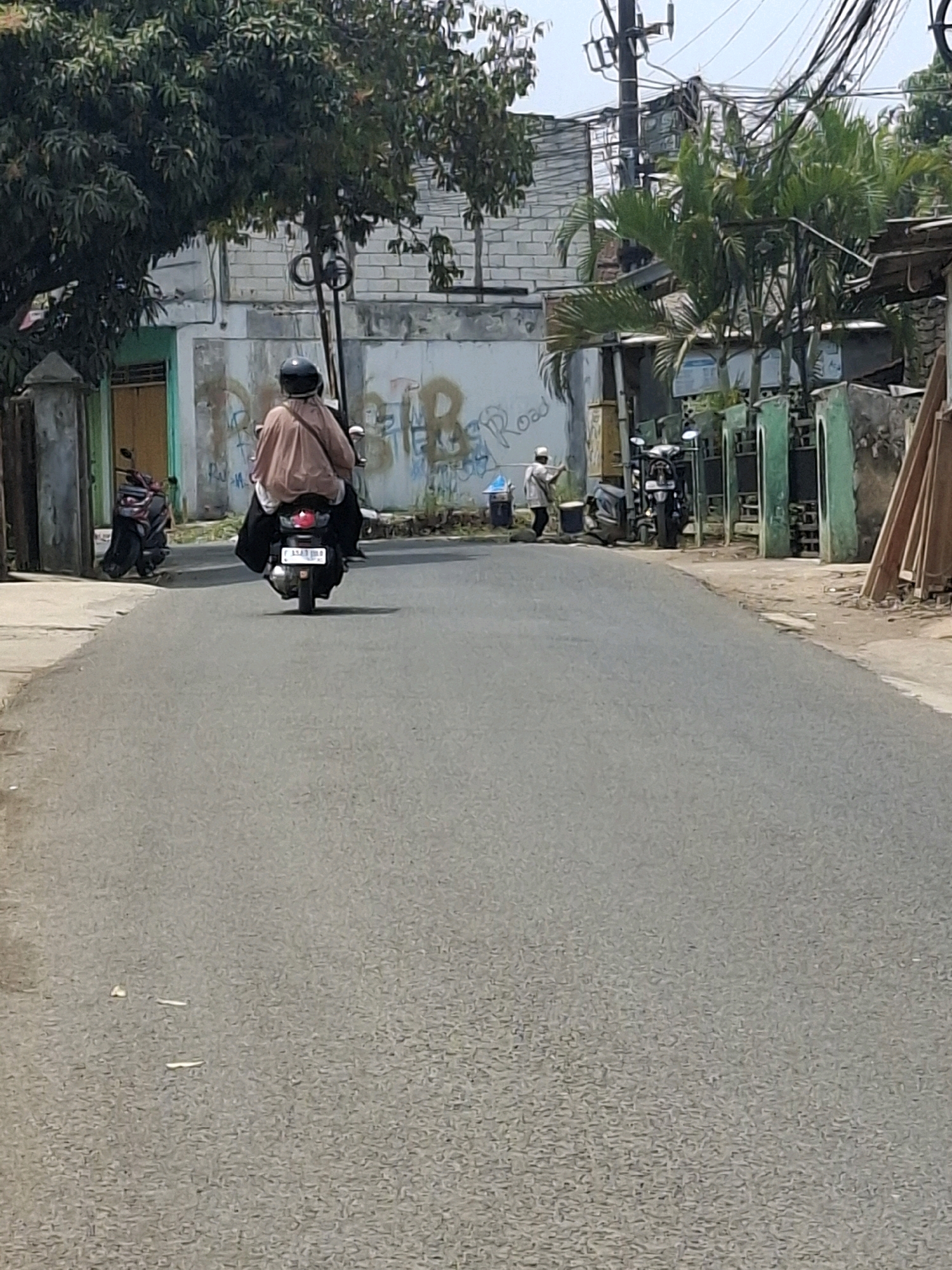 A person wearing a helmet and a light brown robe rides a scooter down a paved road in a residential area of Ciheulang Tonggoh, Indonesia. The road is bordered by buildings with white brick walls, some with graffiti, and fences. There are trees and palm trees along the side of the road. Another person, wearing a white cap and robe, stands on the side of the road near a trash can and a parked scooter. The scene appears to be during daylight hours.