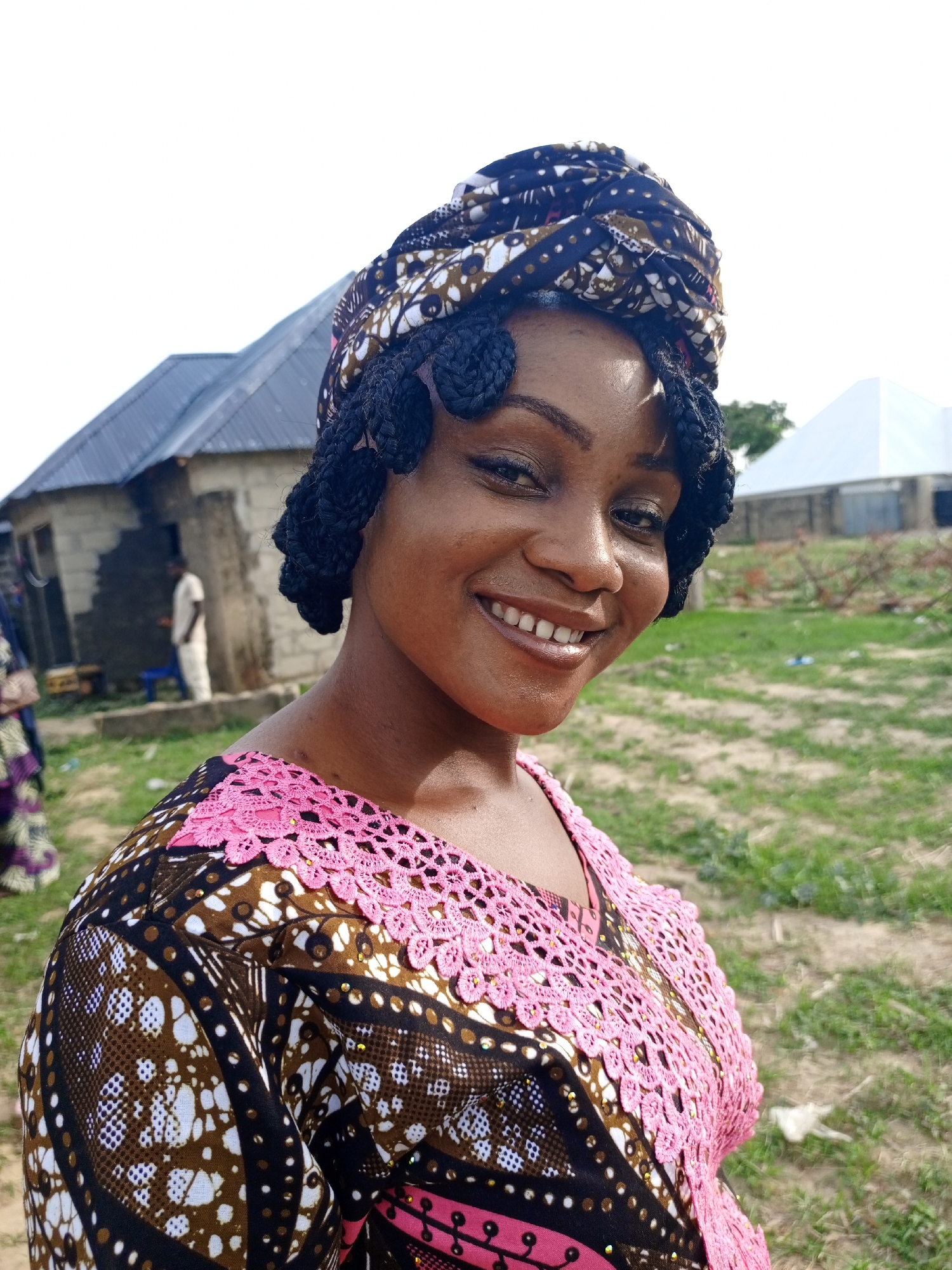 A woman stands smiling at the camera in an outdoor setting, possibly in Keffi, Nigeria, judging by the background structures. She is wearing a headwrap and a patterned top with pink lace detailing around the neckline. Behind her are structures including a house and a tent or temporary building, and scattered vegetation or grassy ground. A figure in white can be seen near the house.