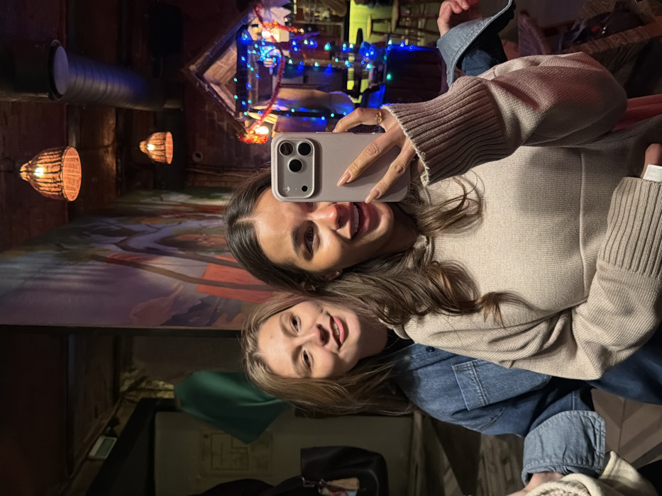 Inside the Bar & Grill "BULGAKOV" (Булгаков) located at Mira Avenue, 19, Krasnoyarsk, Russia, two young adult females are depicted in a mirror selfie. The female in the foreground (right) has long, dark brown hair and wears a light beige hooded sweatshirt with ribbed cuffs. She holds a light-colored smartphone with a triple-lens camera array in her right hand, angled to capture their reflection. Her face shows a wide smile or laugh, exposing teeth, and a small ring is visible on her right ring finger. The second female (left), with light brown hair, stands behind and slightly to the left of the first, wearing a blue denim jacket. She looks towards the camera with a subtle smile.
The background features a large, colorful mural with abstract or stylized forms on the left wall. The ceiling displays exposed wooden beams and silver ductwork, alongside multiple decorative pendant lamps with woven or wicker shades, emitting warm light. To the right, a distinctive small house-like structure, possibly a bar area or booth, is festively adorned with numerous blue, red, and yellow string lights. Exposed brickwork is visible behind this structure. A dark area with a hanging green garment and a partial view of what appears to be a coat rack is present in the far left background. The overall lighting is warm and subdued, indicating an indoor evening or night setting.