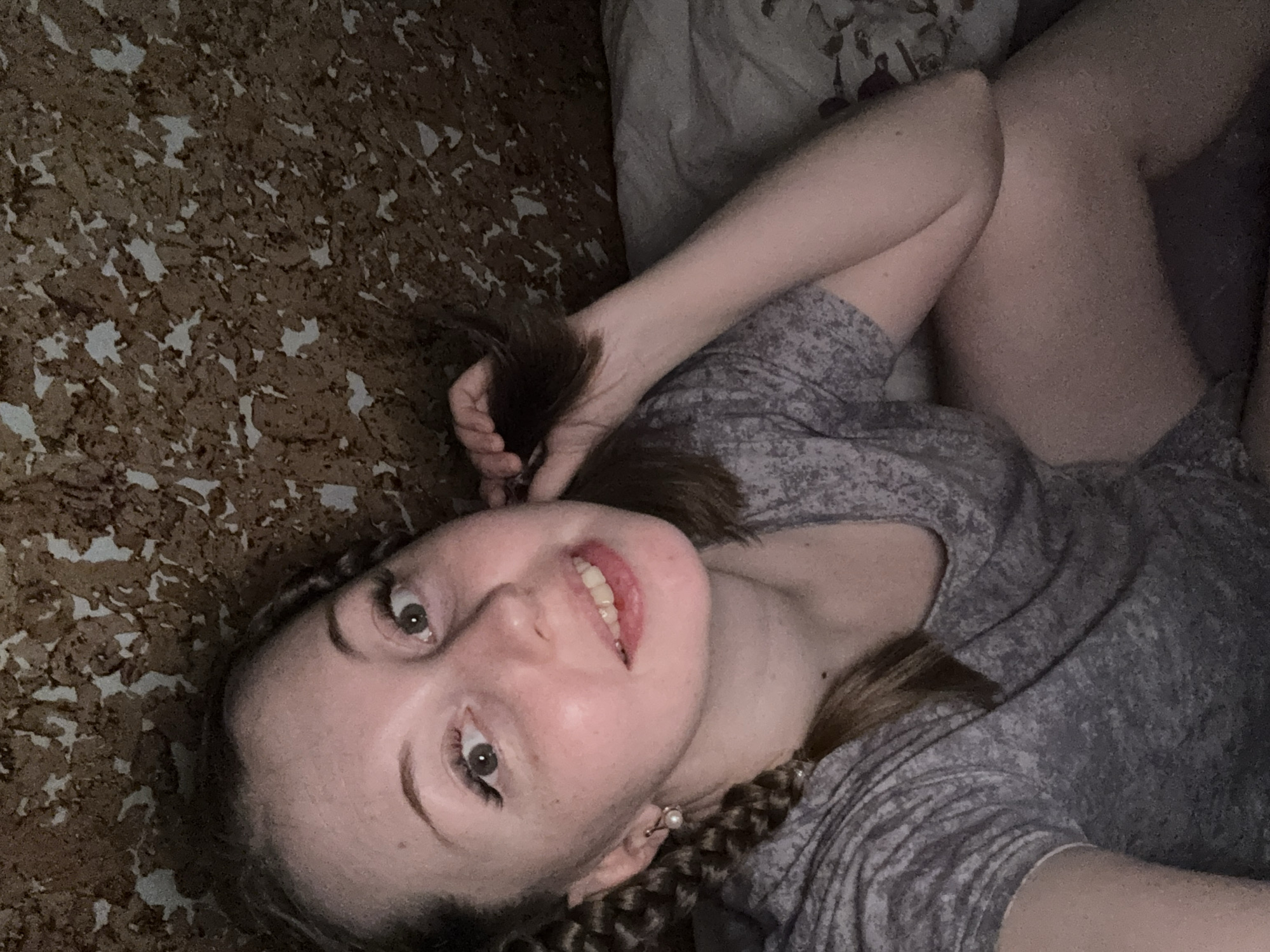 An image taken from an overhead perspective shows a young woman lying down indoors, looking up towards the camera with a smile. She has light skin, dark hair, and light-colored eyes. Her hair is styled with braids on at least one side of her head, and she is wearing a light-colored pearl earring. She is wearing a grey t-shirt with a subtle pattern. Her right arm is bent, and her hand is resting near her head, holding a section of her hair. Her legs are also visible, bent at the knees, and she appears to be wearing shorts or underwear that match the grey top.
The background consists of two main elements. To the left and behind her head, there is a patterned surface that resembles cork board or a textured wall, with irregular brown and beige patches. To her right and beneath her body, there is a grey fabric surface, likely a bed, couch, or blanket, possibly with a subtle pattern. The lighting is somewhat dim and soft, suggesting an indoor setting without direct harsh light. Her expression is pleasant and engaging. There is no visible text in the image.