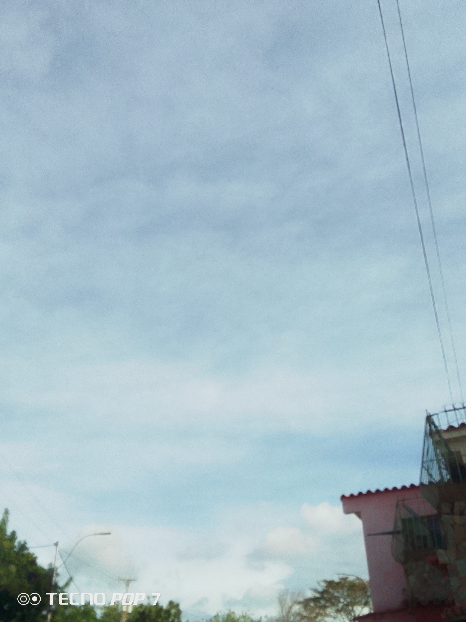 The image shows a cloudy sky with two electrical wires running vertically on the right side. In the lower right corner, there is a portion of a pink building with a red tiled roof and a wire mesh structure. In the lower left corner, green trees and a street light are visible. The camera model "TECNO POP 7" is displayed in the bottom left corner. The location is Altagracia, Venezuela.