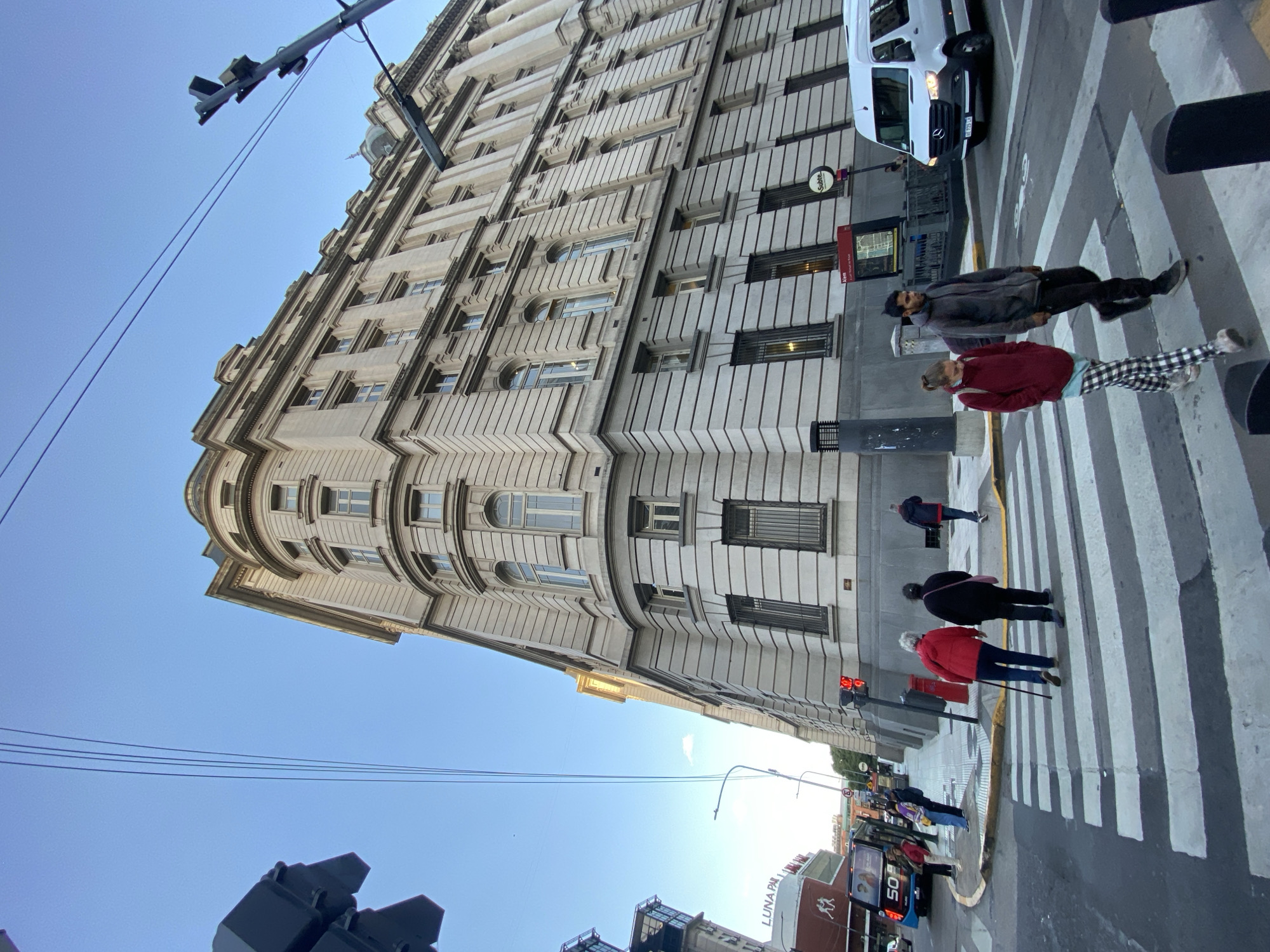 The image depicts a street scene in Buenos Aires, Argentina. A tall, ornate building with multiple windows dominates the left side of the frame, partially angled towards the sky. A pedestrian crossing is visible, with several people walking across it. A white van is parked on the right side of the street. Streetlights and traffic lights are present, adding to the urban environment. A bus with "LUNA PARK" written on it can be seen in the background.