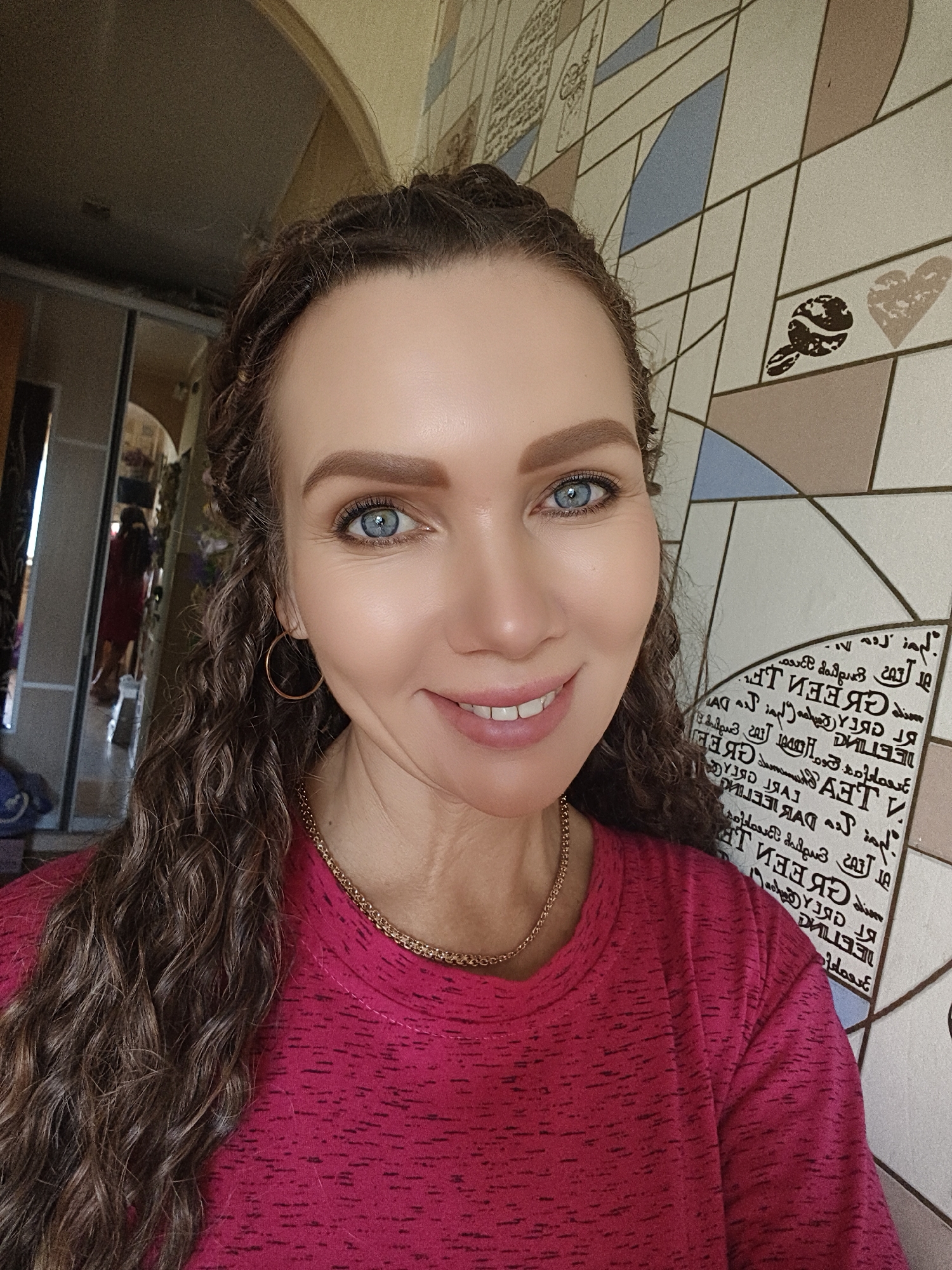 A woman with blue eyes and long, curly brown hair is pictured in a close-up, front-facing shot. She is wearing a pink t-shirt and a gold chain necklace. Her hair is styled in braids that frame her face. The background consists of a tiled wall with a geometric pattern and some decorative elements. The lighting suggests an indoor setting, possibly a home. There are no other individuals visible in the immediate foreground. The image is a selfie taken from a slightly low angle.