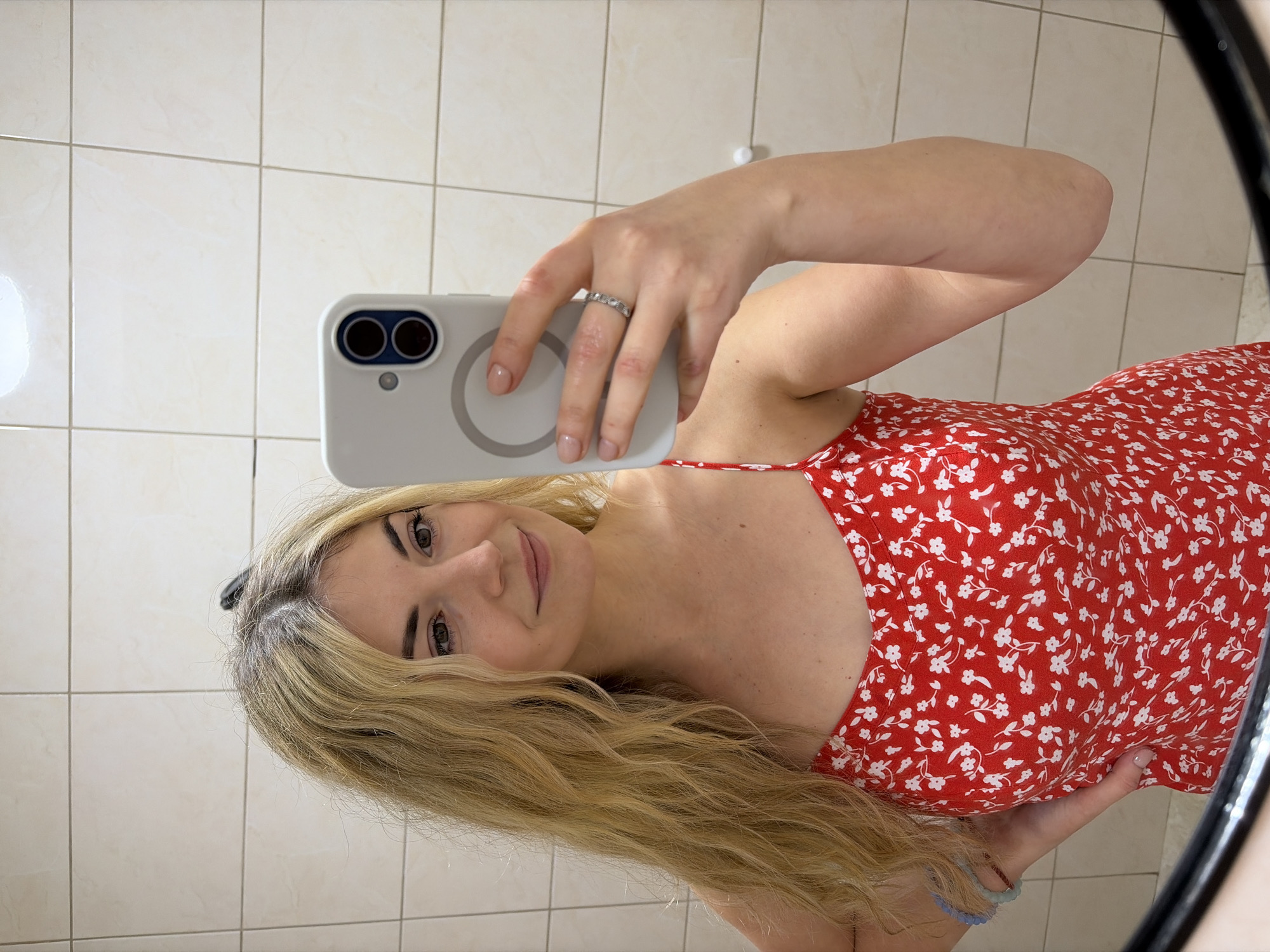 A female subject, appearing to be a young adult with long, wavy blonde hair exhibiting darker roots, is captured in a mirror selfie within an interior tiled space. She wears a red sleeveless dress patterned with small white flowers and leaves. Her right hand holds a light gray smartphone, visible with a dual camera system and a MagSafe ring on its back. A silver-colored ring with small decorative elements is present on her right ring finger. Her left hand rests on her hip, revealing light blue and brown beaded bracelets on her left wrist. The background is composed of uniform light beige square ceramic tiles with dark grout lines. This scene is located within an interior tiled space in Budva, Montenegro.