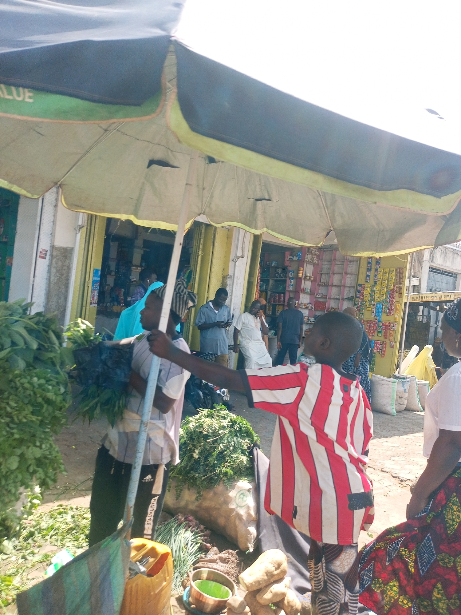 The image depicts a bustling outdoor market scene under the shade of a large, dark green and light green umbrella. Bright daylight illuminates the area, casting strong light.
In the foreground to the right, a person wearing a red and white vertical striped shirt stands with their right arm extended. To the left of the frame, another person, dressed in a striped shirt and dark trousers with "MALISTYLE" written on the leg, holds a vertical pole, likely supporting the umbrella. They are surrounded by large bundles of fresh green leafy vegetables. Near their feet, a yellow jerrycan, a green bucket, and several pieces of produce resembling yams or potatoes are visible on the dusty ground.
Various other people are present in the mid-ground and background, including a woman in a patterned skirt to the far right. Several individuals are seen walking or standing near storefronts that line the street behind the market stalls. These storefronts have yellow roll-down doors and shelves stocked with various items, including packaged goods and cans. To the far right in the background, white sacks or bags are stacked on the ground. A sign with partially visible text, possibly reading "HAWA MASIID SUYA," is seen on one of the background buildings. The atmosphere appears lively and functional, consistent with an active market day.