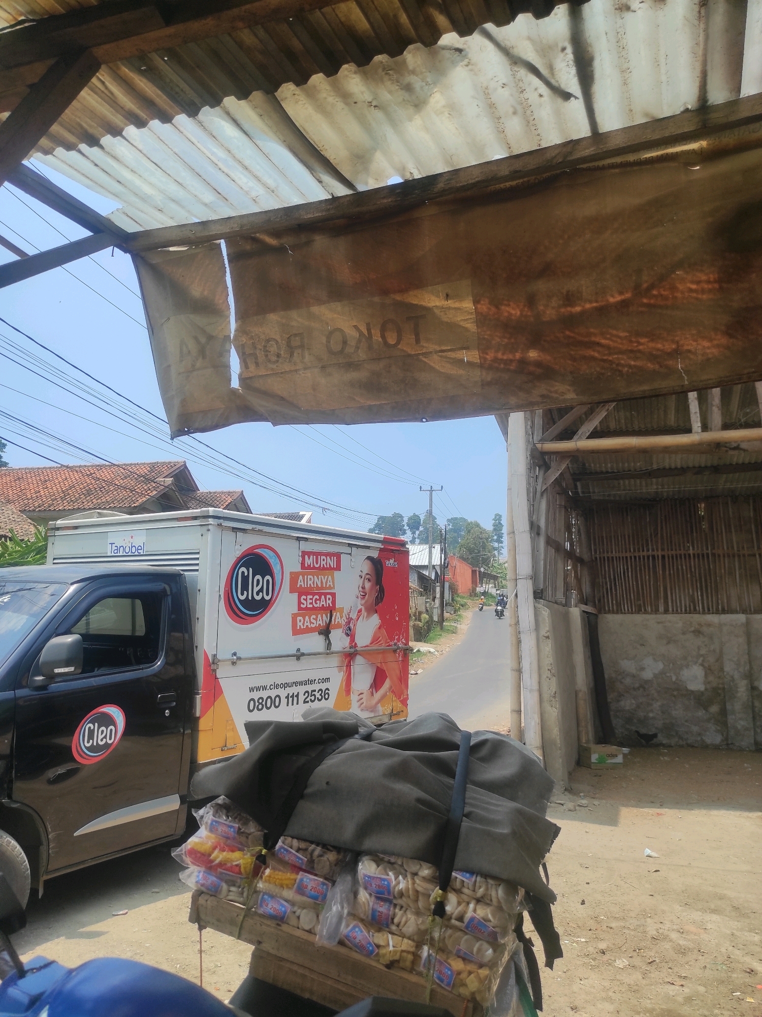 This image depicts a street scene in Pasirlangu, Indonesia. In the foreground, a motorcycle is partially visible, with a stack of goods wrapped in dark fabric on its rear. These goods appear to be individual packages of snacks or food items, many of which have blue and red labels with text that includes "2000."
To the left of the motorcycle is the side of a black pickup truck. The truck has a logo on its door that says "Cleo." Behind the truck is a white delivery truck with branding that includes "Tanubel" on its side and a large circular "Cleo" logo with an image of a woman. Text on the truck reads "MURNI AIRNYA SEGAR RASANYA" and "www.cleopurewater.com 0800 111 2536."
The background shows a rural or semi-urban environment. A paved road runs past the vehicles, with houses and vegetation visible on either side. A power pole with overhead wires is present on the left. Further back, trees and buildings are scattered across the landscape.
Above the scene, a makeshift roof structure is visible, constructed from corrugated metal and fabric. The sky is clear and blue. The overall impression is of a commercial activity or delivery taking place outdoors.