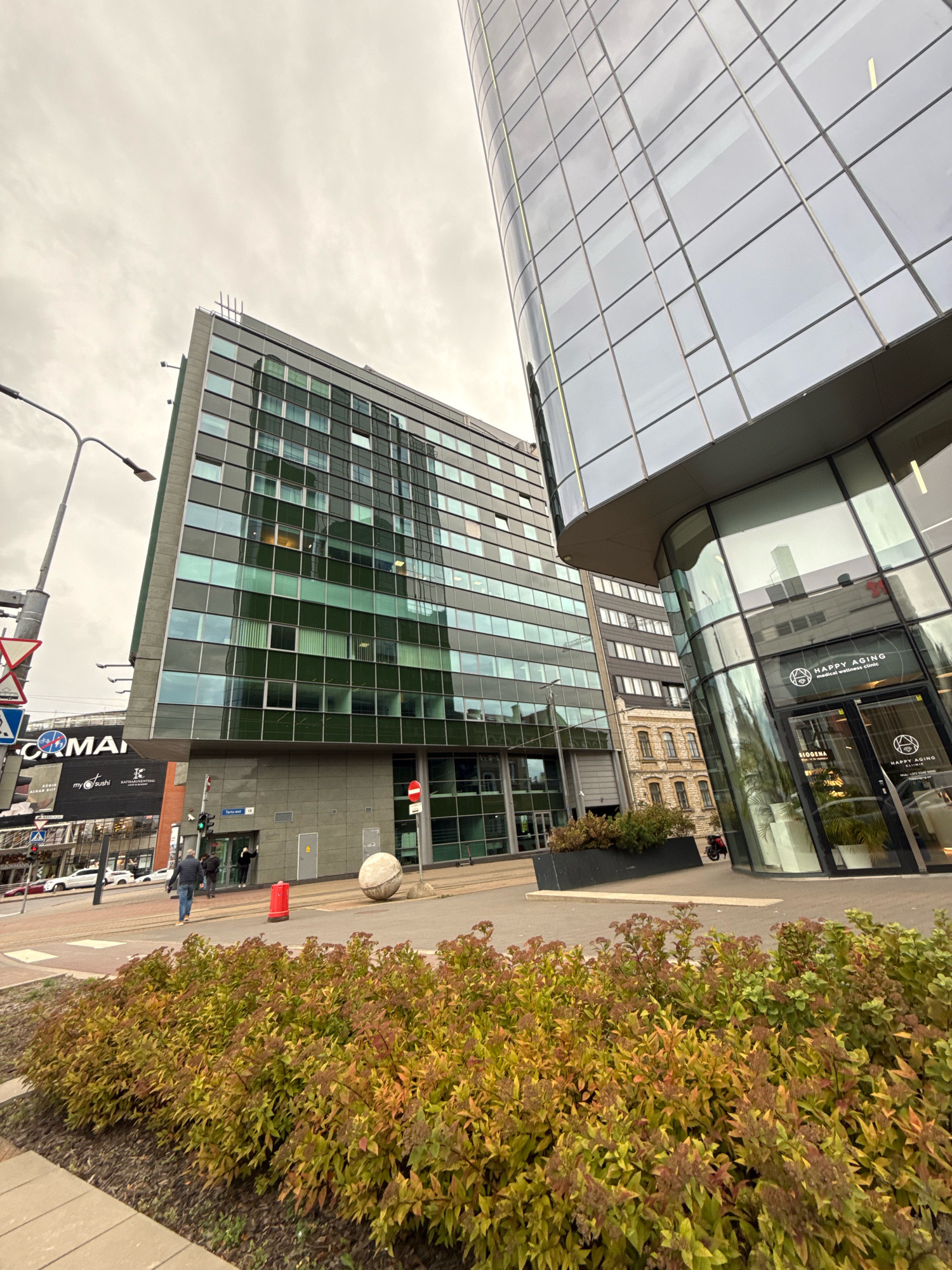 The image shows a street view in Tallinn, Estonia, focusing on modern architecture. In the foreground, a low-lying bush with autumnal foliage is visible. Behind it, a large, multi-story building with a green-tinted glass facade dominates the left side of the frame. This building features numerous rectangular windows, some reflecting the cloudy sky. To the right, a newer, taller building with a curved, glass facade curves into the frame. The glass panels are large and reflective, showing glimpses of interior spaces and the sky. Signs are visible on the buildings, including one for "HAPPY AGING medical wellness clinic" on the curved building's entrance. A street with traffic lights and road signs is present in the mid-ground. Two individuals are seen walking on the sidewalk in front of the green-facade building. A large, spherical stone object is situated on the street near the sidewalk. The sky is overcast, contributing to the muted lighting of the scene.