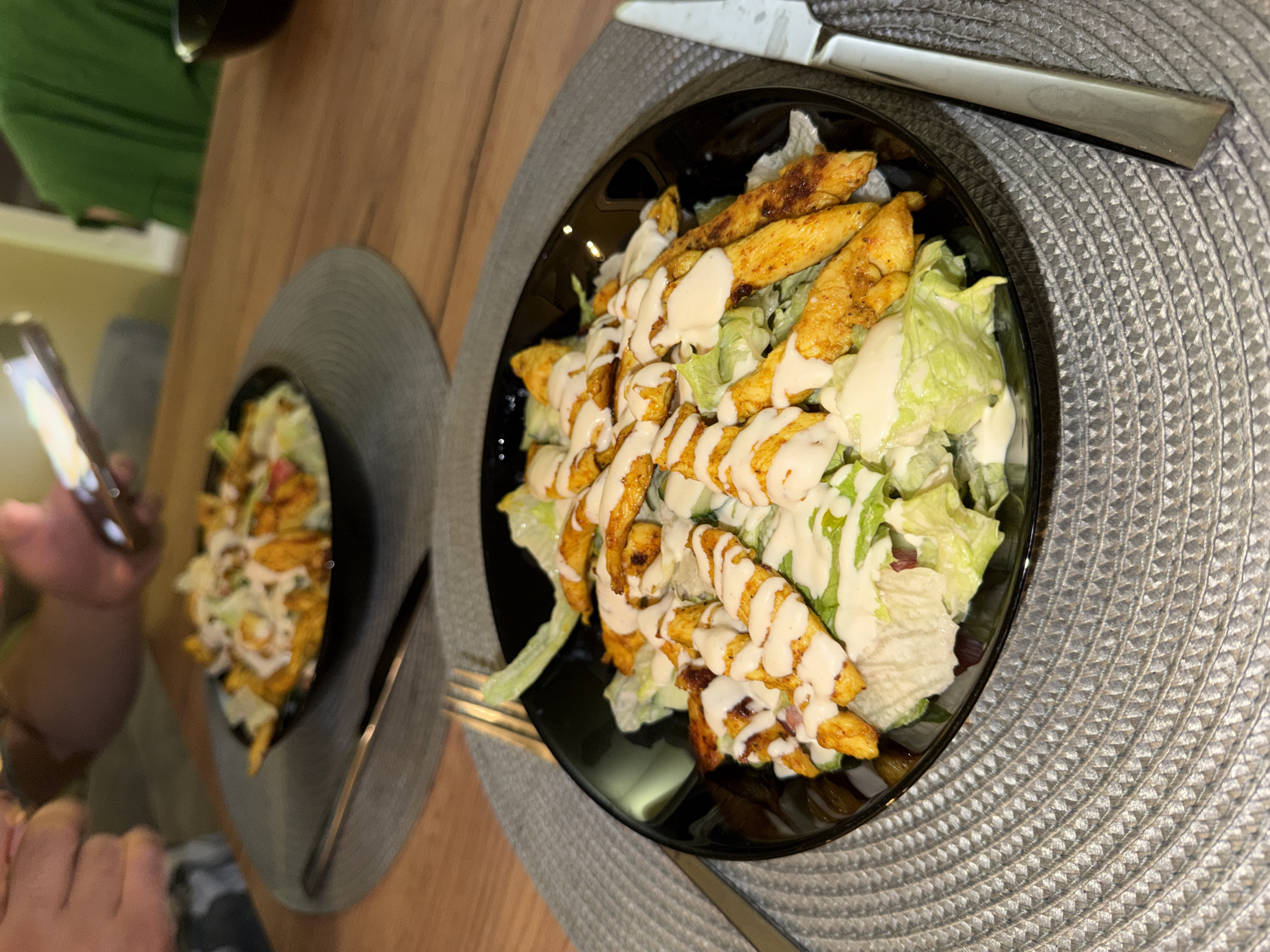 Two black bowls of salad with grilled chicken strips and white sauce are presented on a wooden table. The salad in the foreground is closer to the camera and features a generous portion of lettuce topped with seasoned chicken strips and a creamy dressing. Utensils, a fork and a knife, are placed on the table next to the bowl. In the background, another similar bowl of salad is visible, and two individuals are partially seen, one holding a smartphone. The setting appears to be an indoor dining area, possibly a restaurant or home in Varna, Bulgaria.