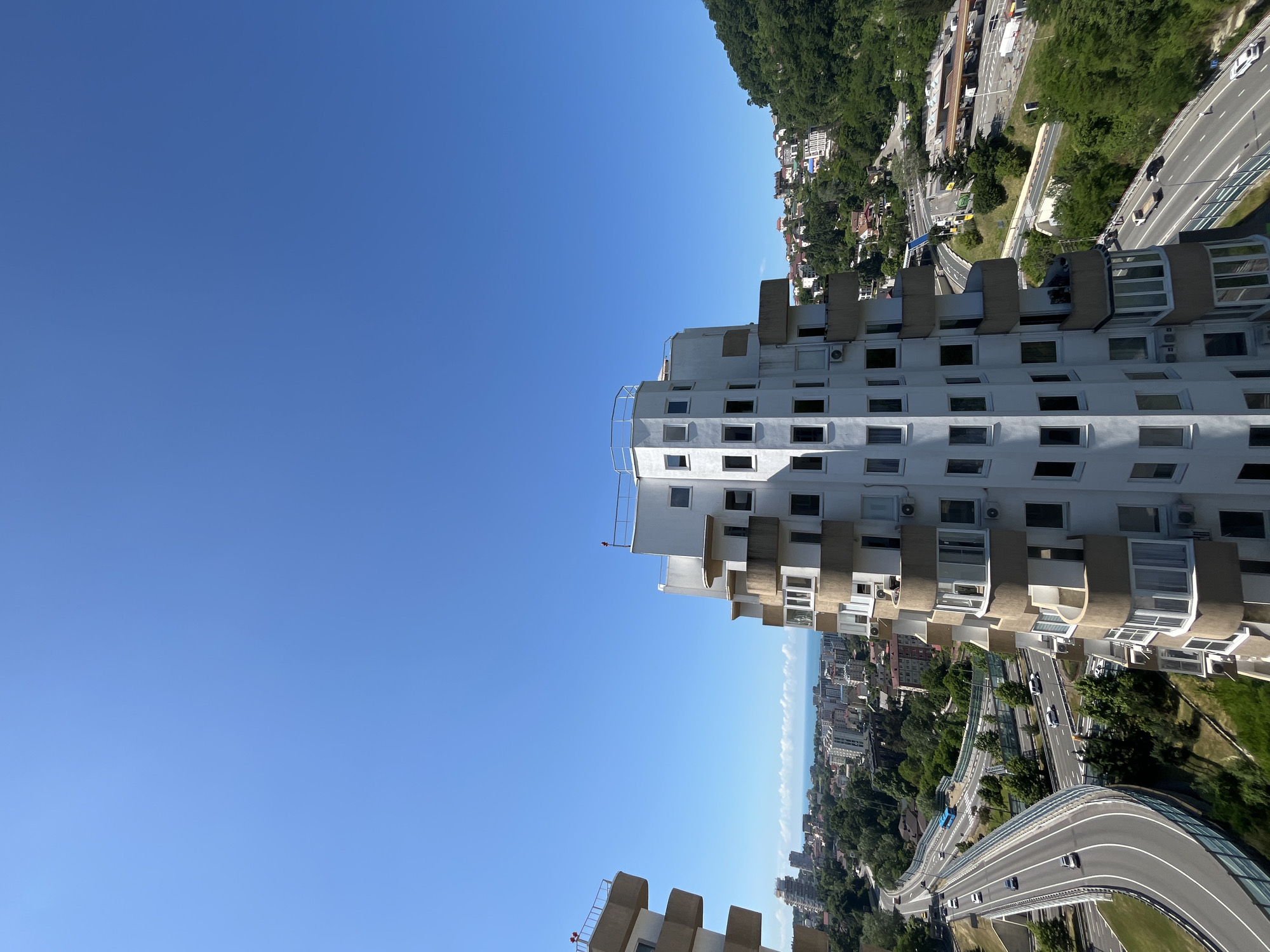 An aerial view captures a segment of Sochi, Russia, under a clear blue sky. In the foreground, a modern, white apartment building with a distinctive stepped, triangular facade dominates the right side of the frame. The building features numerous windows and protruding balconies with curved, beige railings. To the left, a portion of another similar apartment building is visible. Below and to the left, a multi-lane highway curves through a landscape dotted with trees and other buildings. Cars are present on the highway, and a blue bus is also visible. The background reveals a coastal city with more buildings extending towards a hazy horizon where the sea meets the sky. A lush, green hillside rises to the right of the city, also populated with houses. The scene appears to be taken on a bright, sunny day.