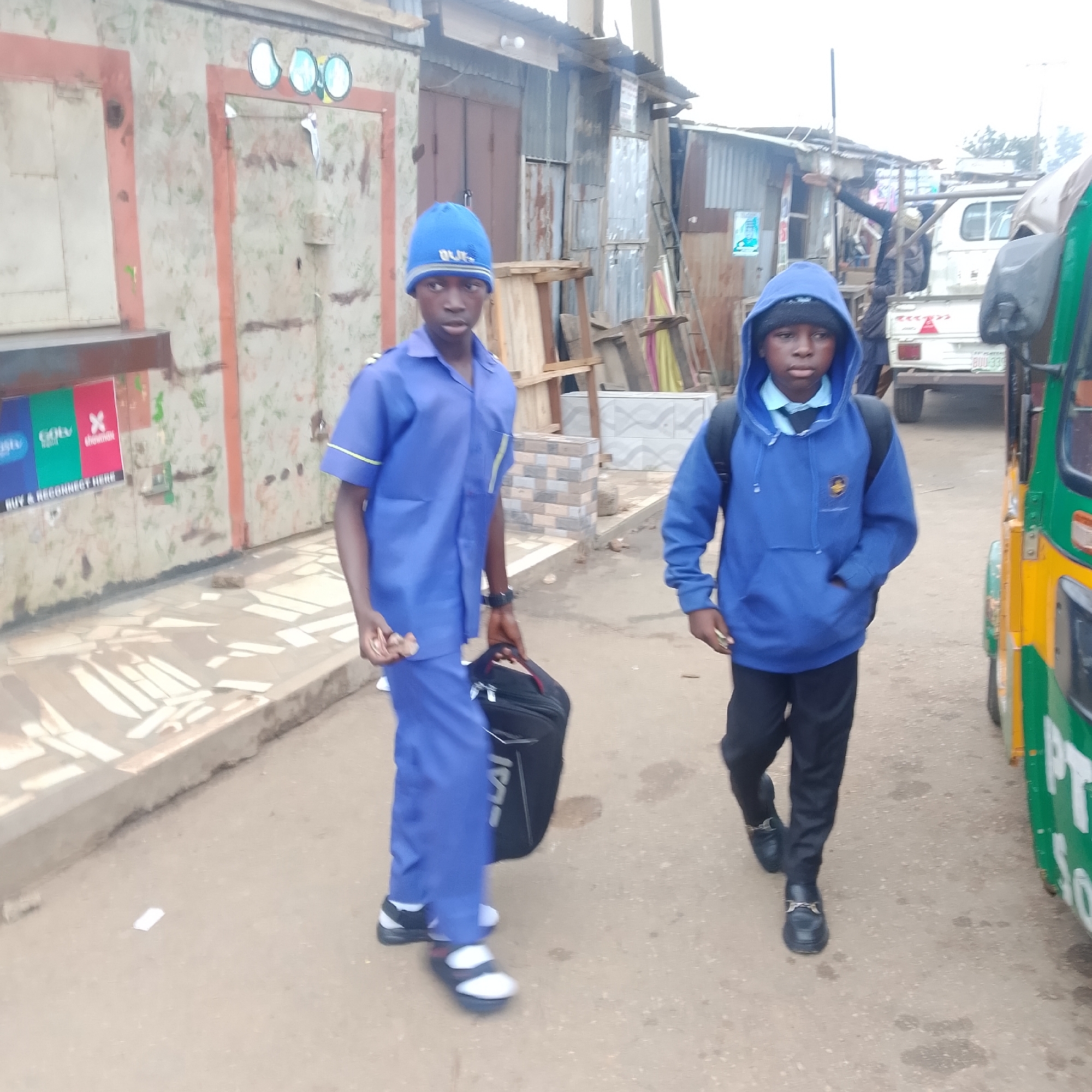 Two young boys are walking on a dirt road in front of a weathered, stall-like structure. The boy on the left wears a blue uniform with a yellow trim and a blue beanie with "BULS" written on it. He is carrying a black bag in his right hand and wears white socks with black and white sandals. The boy on the right wears a blue hooded sweatshirt over a blue collared shirt and tie, and black pants with black loafers. A yellow and green vehicle, possibly a tuk-tuk, is visible on the right side of the image. The environment appears to be a market or street in a developing area, with corrugated metal and aged buildings in the background.