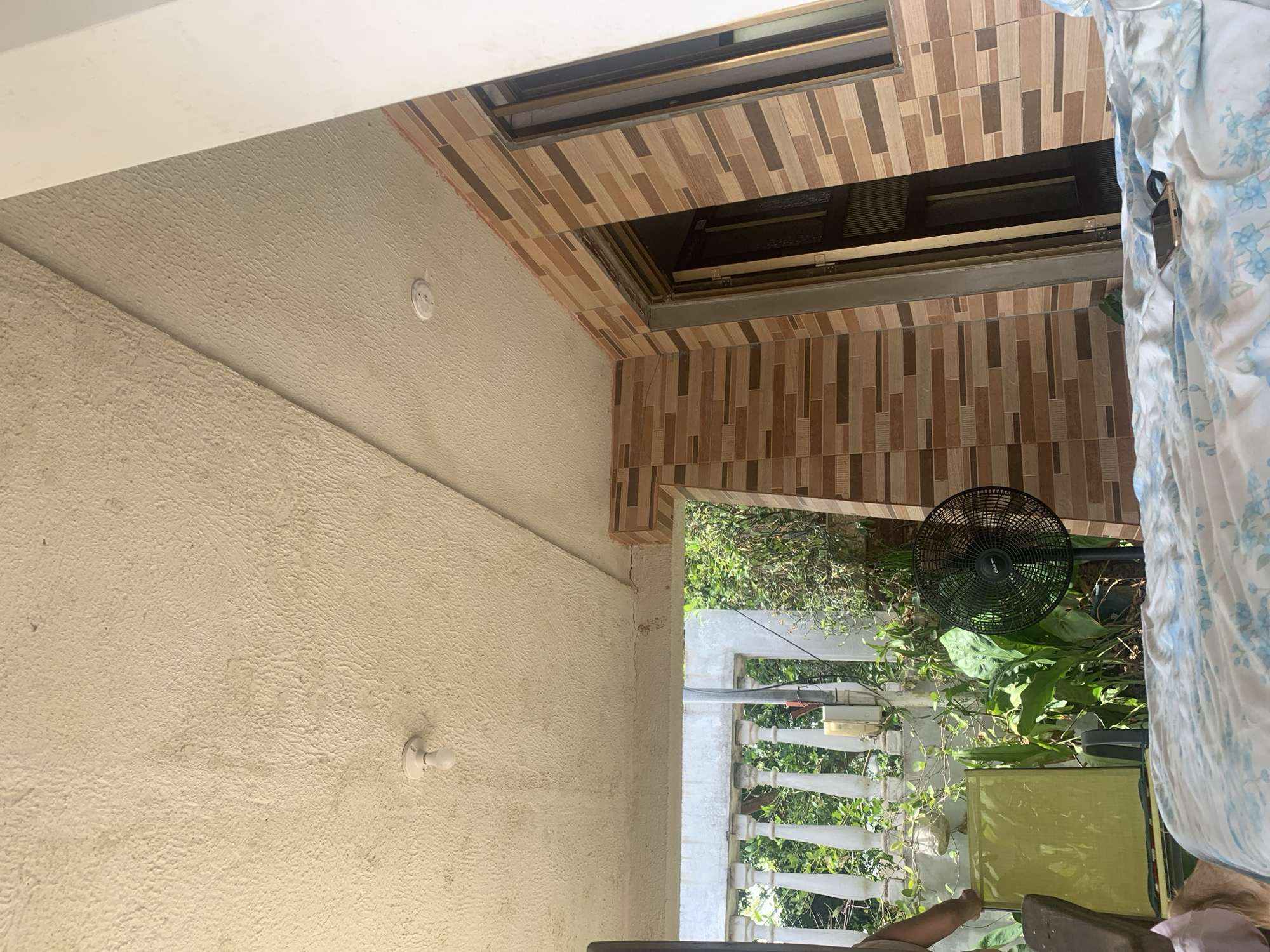 The scene depicts a covered outdoor living space, likely a porch or patio, located in Santa Ana, Venezuela, during daylight hours under clear skies. The perspective is from a low vantage point, looking across a bed or seating area towards the exterior.
The overhead structure features a light beige, textured ceiling with two light fixtures: one exposed bare bulb fixture in the foreground and one recessed light fixture further back. The ceiling exhibits minor discoloration. To the right, a wall is clad with horizontal rectangular tiles in varying shades of brown and beige, arranged in a staggered pattern. This tiled wall contains an open doorway with a partially visible dark-colored door and a window with dark framing. A thick white column partially obstructs the far right.
In the mid-ground, a white balustrade with several balusters defines the edge of the covered area, beyond which is an exterior view of dense green foliage and trees. A utility box or electrical component is mounted on a slender post behind the balustrade, with associated wiring visible.
In the lower left, the upper arm and shoulder of an individual wearing a light-colored garment are partially visible, positioned near a bright yellow-green folding chair or recliner. Adjacent to this, a black stand fan with a circular cage is situated on the ground, surrounded by several green potted plants. The immediate foreground is covered by a blue and white floral-patterned fabric, upon which a dark-colored smartphone and a smaller black accessory are resting.