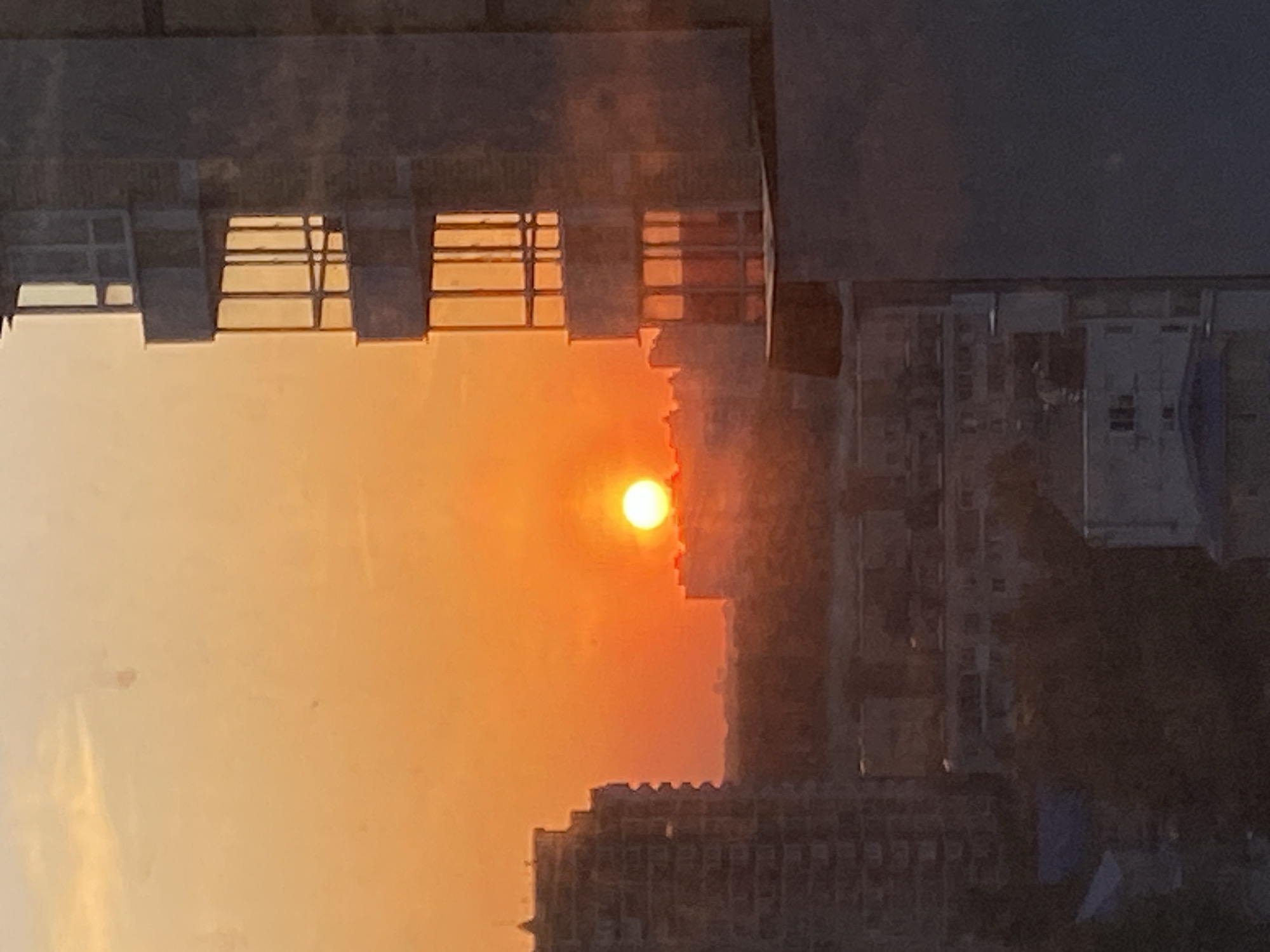 The image shows a sunset in a city. The sun is a bright, round orange disc low in the sky. The sky is a gradient of colors, ranging from yellow near the horizon to orange and pink higher up. Silhouettes of buildings are visible in the foreground and background. The image appears to be taken from an elevated position, possibly from inside a building looking out through a window frame. The window frame is visible in the upper parts of the image, appearing as dark, rectangular shapes.