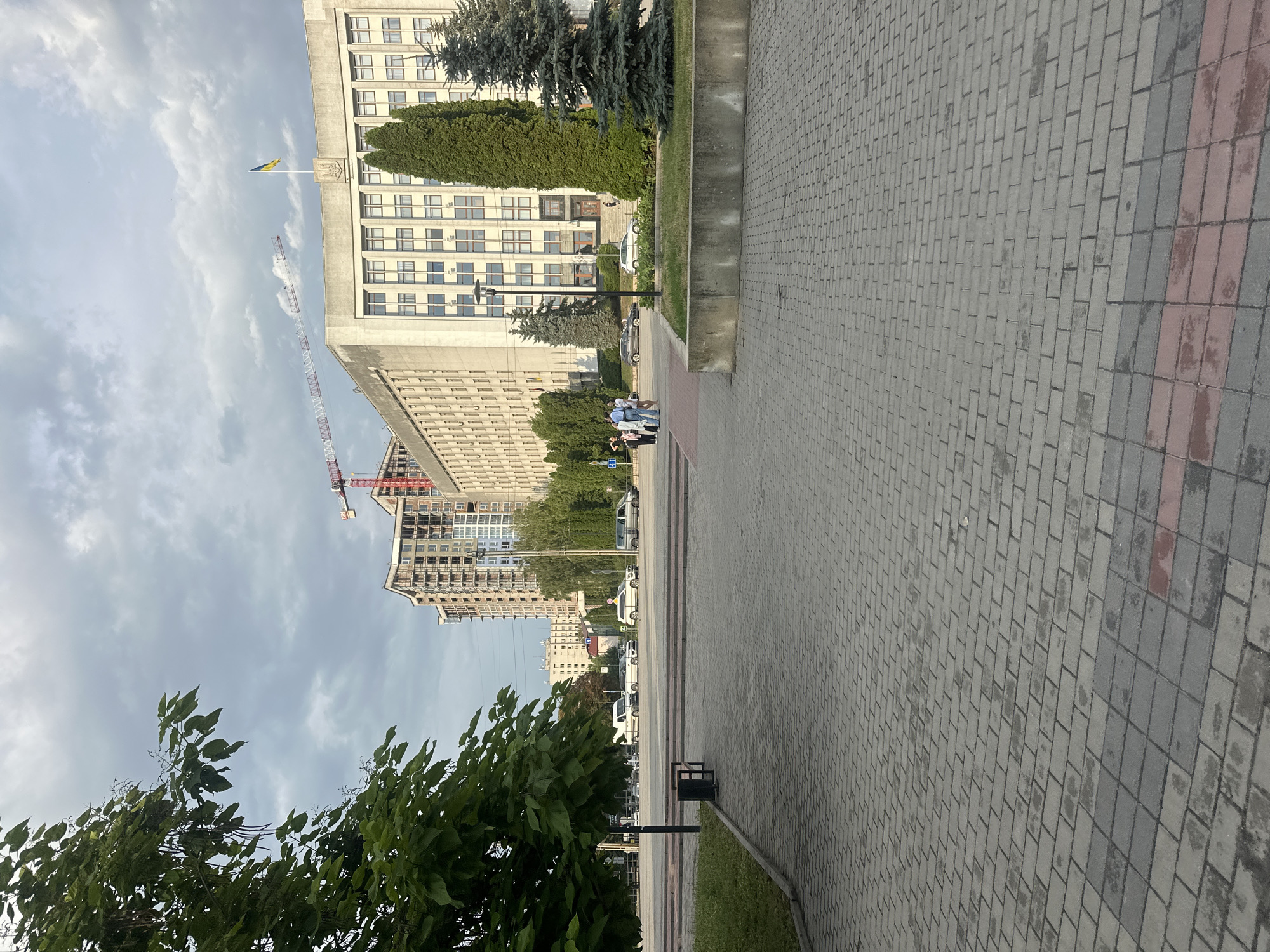 The image depicts a street scene in Ternopil, Ukraine, characterized by a large administrative-style building with multiple stories and numerous windows. A Ukrainian flag flies from a pole on the building's facade, above an emblem. Construction is evident, with a red and white crane towering over a modern apartment building under construction in the mid-ground. The foreground features a wide, paved walkway made of interlocking bricks in shades of gray and red. Lush green trees and foliage border the walkway and extend into the scene.
Several cars, including vans and what appears to be a white sedan, are parked along the street. A small group of people, likely pedestrians, are visible on the sidewalk near the trees in the lower left portion of the image. A trash receptacle stands near the edge of the walkway. The sky is overcast with dramatic clouds, suggesting either early morning or late afternoon, with diffused daylight. The overall atmosphere is one of urban activity with elements of construction and civic presence, under a moderately cloudy sky. There is no visible text in the image.