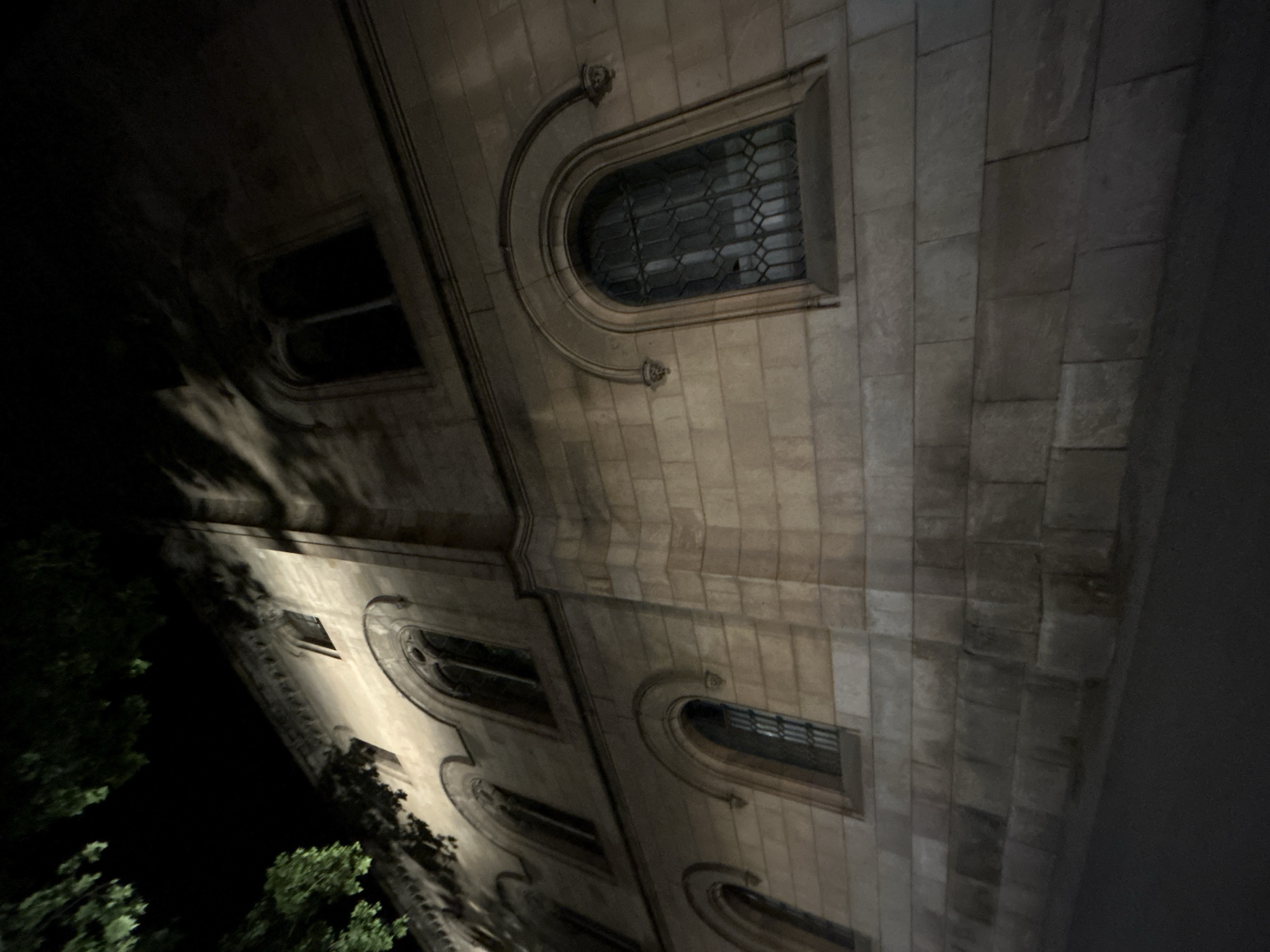 A low-angle shot shows the exterior of a stone building at night. The building features a series of arched windows, some with decorative ironwork visible through the glass. The facade is composed of stone blocks, with visible texture and aging. The lighting suggests a dark environment, possibly a city street. Trees are visible in the lower left corner, hinting at vegetation in the vicinity. The location is Barcelona, Spain, as implied by the presence of specific architectural styles common in the city.