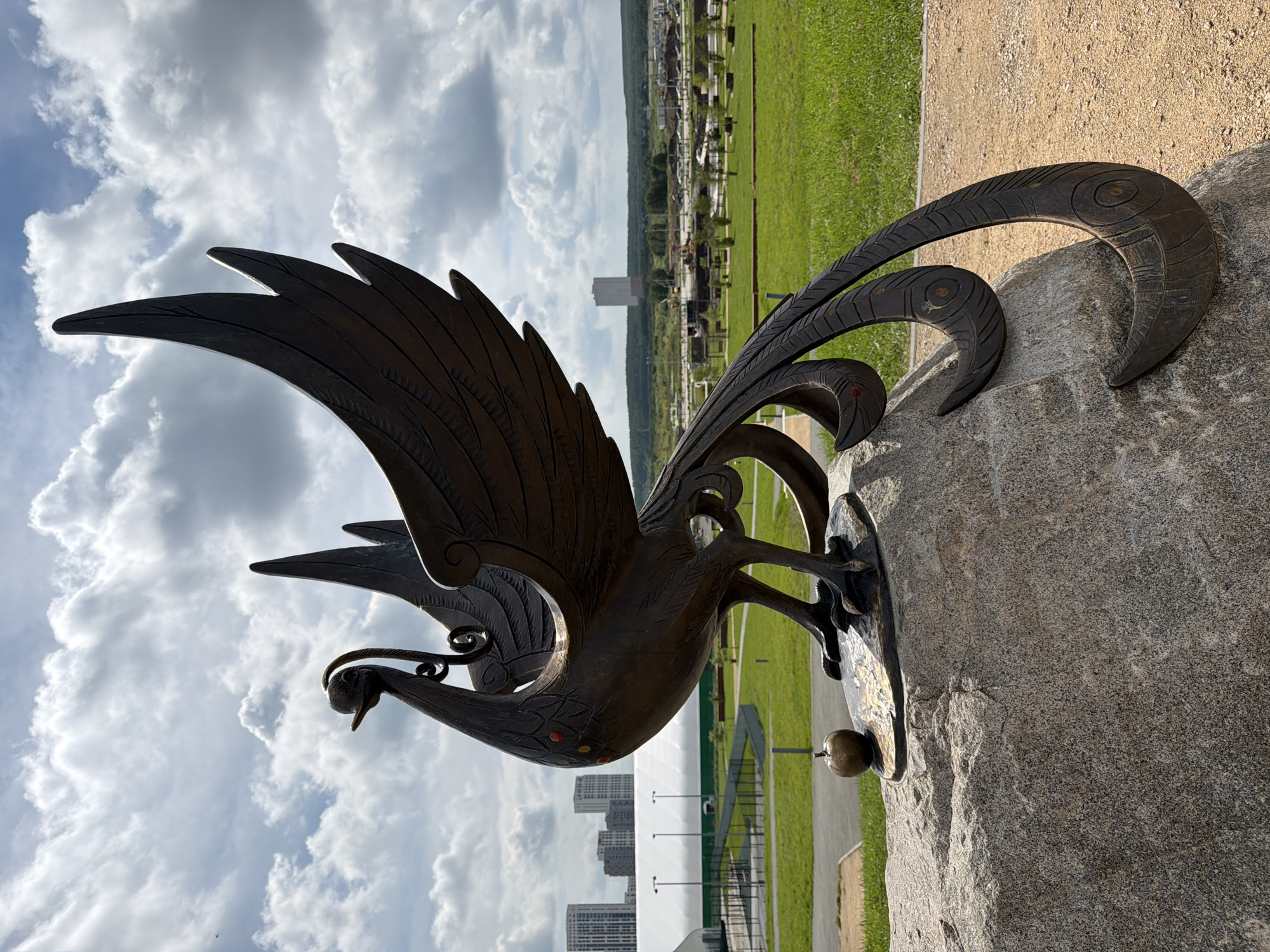 The image depicts a bronze sculpture of a mythical bird, likely a phoenix, positioned atop a large, rough-hewn stone. The sculpture is dark brown and detailed, featuring intricate feather patterns on its wings and tail. The bird has a slender neck, a beaked head with a decorative element above it, and sharp talons gripping a small apple. The sculpture is set in an outdoor park environment. In the background, there is a vast expanse of green grass, a walking path, and several modern buildings in the distance. The sky is filled with dramatic, heavy clouds, suggesting a dynamic weather condition.
This sculpture is located in Yekaterinburg, Russia, specifically within the "Yeltsin Center Park" or "Yeltsin Center Sculpture Park." The park is part of the broader Yeltsin Center complex, which also houses a museum and cultural center dedicated to Boris Yeltsin. The architectural style of the distant buildings, particularly the taller ones, is consistent with modern urban development in Yekaterinburg. The sculpture itself is known as "The Phoenix" and is a prominent piece within the park's collection of contemporary art.