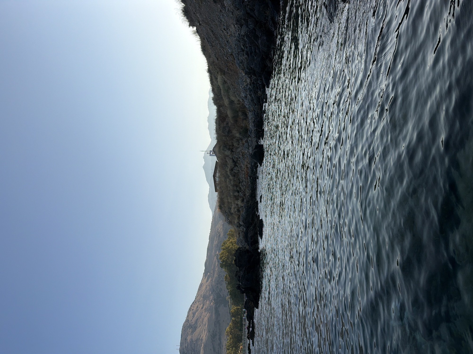 This is a photograph taken from the water, looking towards the shore of Georgioupoli, Greece. The foreground is dominated by the sea, with gentle ripples creating a textured surface of dark blue and green hues. The water appears clear, with some darker areas indicating greater depth.
The shore is rocky and somewhat rugged. A cluster of dark, jagged rocks is prominent in the mid-ground, jutting out into the water. Beyond the rocks, the land slopes upward, covered in dry vegetation and scattered rocks. On the crest of the hill, a small, white building with a cross on top is visible, possibly a chapel. Further in the distance, a larger, flat-roofed structure with a darker exterior is present. Behind the immediate coastline, rolling hills form the backdrop, with a more distant mountain range faintly visible on the horizon. The sky is clear and a pale, uniform blue, suggesting it is either early morning or late afternoon. There are no people or animals clearly visible in the frame.