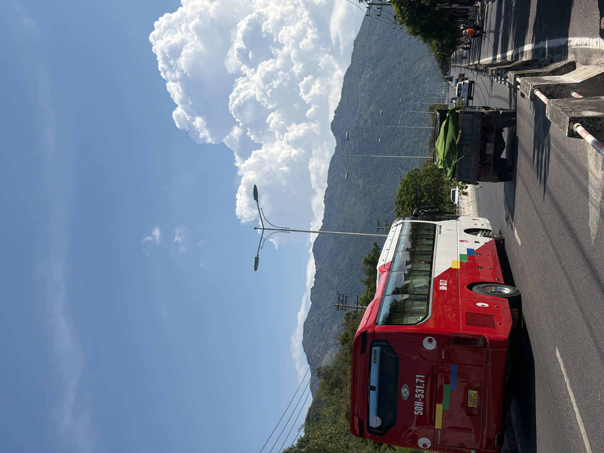A red and white bus is parked on the right side of a highway in Nam Nha Trang Ward, Vietnam, with its rear facing the camera. The bus has a license plate that reads "50H-531.71." A dark green truck carrying a large load of green vegetation is driving in front of the bus. In the background, a lush green mountain range rises under a bright blue sky with scattered white clouds. Several streetlights line the highway, and other vehicles are visible in the distance. The scene appears to be a sunny day.