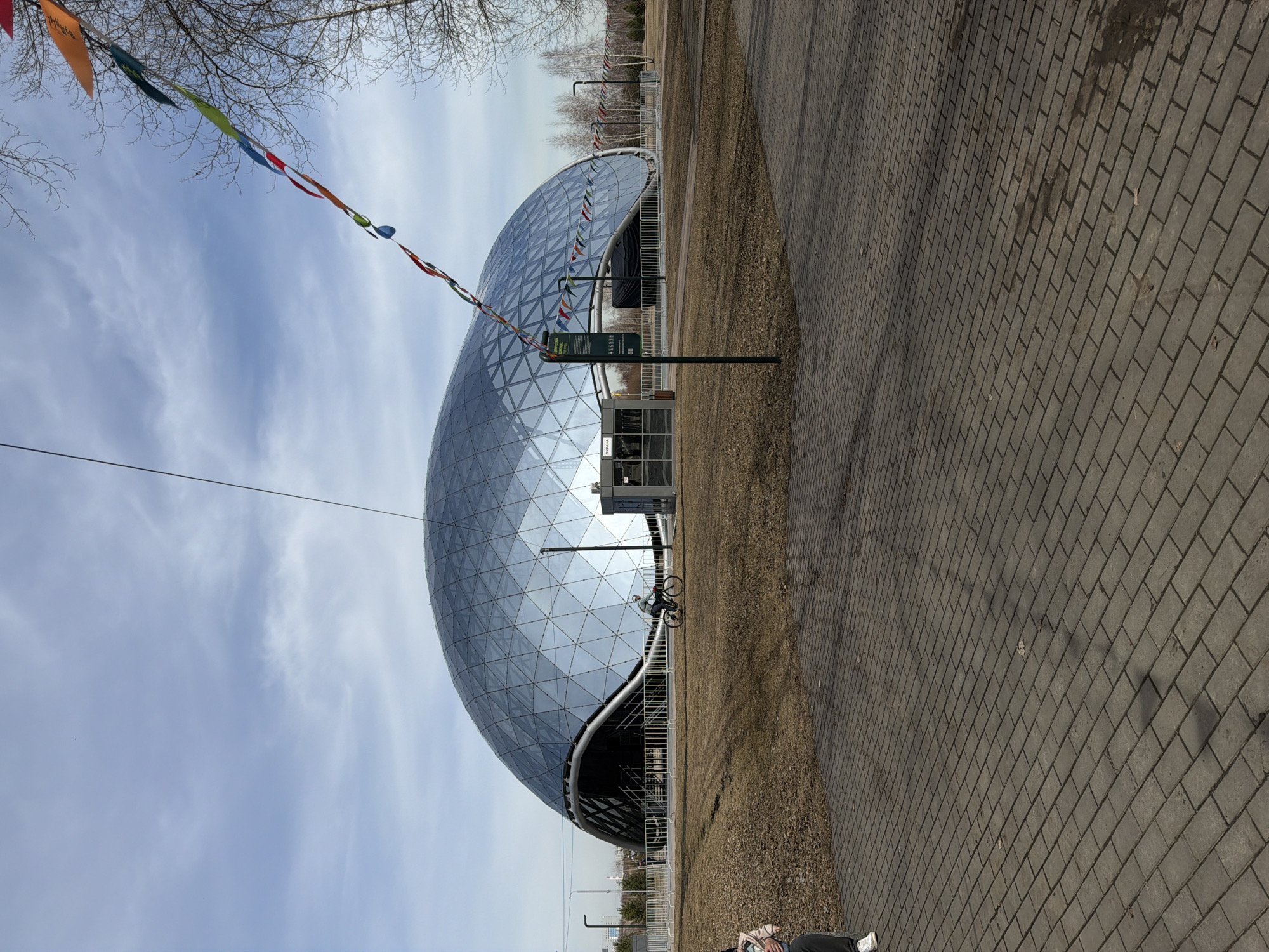 The image shows the “Cube” in Krasnoyarsk, Russia, a distinctive geodesic dome structure. The dome is constructed of triangular glass panels set in a dark frame. A string of colorful pennants extends toward the dome from nearby bare trees. The structure is near a brick-paved road, alongside a narrow strip of land covered with gravel and short grass. A sign stands close to the building entrance. A bicycle and a person in blue clothing can be seen near the dome’s base. The sky is pale blue with streaks of white clouds.