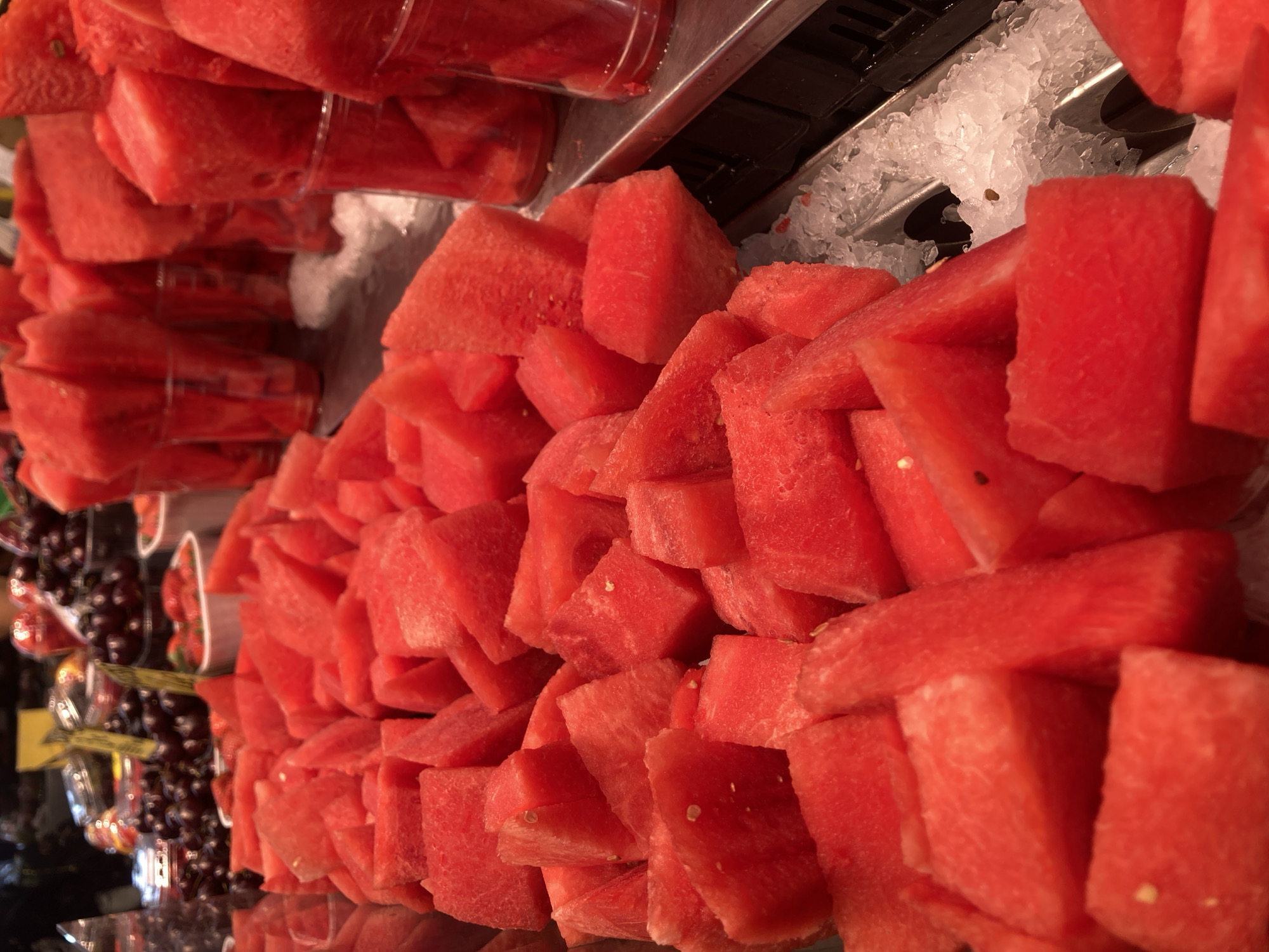 This is a close-up, eye-level shot of a fruit stand, likely in La Boqueria Market in Barcelona, Spain. The foreground is dominated by a large pile of cubed, bright red watermelon pieces displayed on crushed ice. Several clear plastic cups, also filled with watermelon pieces, are visible in the middle ground, suggesting they are pre-portioned servings for sale. In the background, other fruits like grapes and strawberries can be seen in containers, along with some plastic packaging and signage that is out of focus. The lighting appears to be artificial, creating a vibrant and appealing display. There are no people visible in the frame.