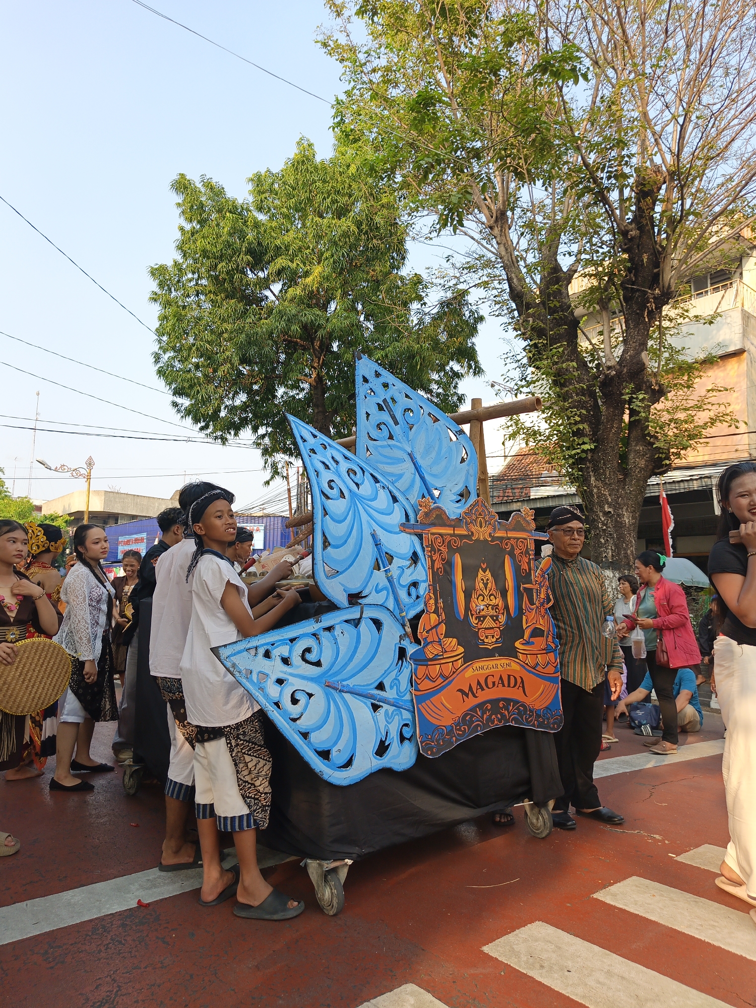 In Klaten, Indonesia, a group of people are participating in a parade or cultural event. The focus is on a large, colorful float decorated with blue, wing-like structures and a central panel featuring an orange and black design with the text "SANGGAR SENI MAGADA." Several individuals, including children and adults, are dressed in traditional attire and appear to be part of the procession, some pushing or standing near the float. The environment is an outdoor urban setting, with trees, buildings, and utility wires visible in the background. The ground is paved with red and white markings, suggesting a street or pedestrian walkway. The lighting indicates it is daytime, possibly late afternoon.