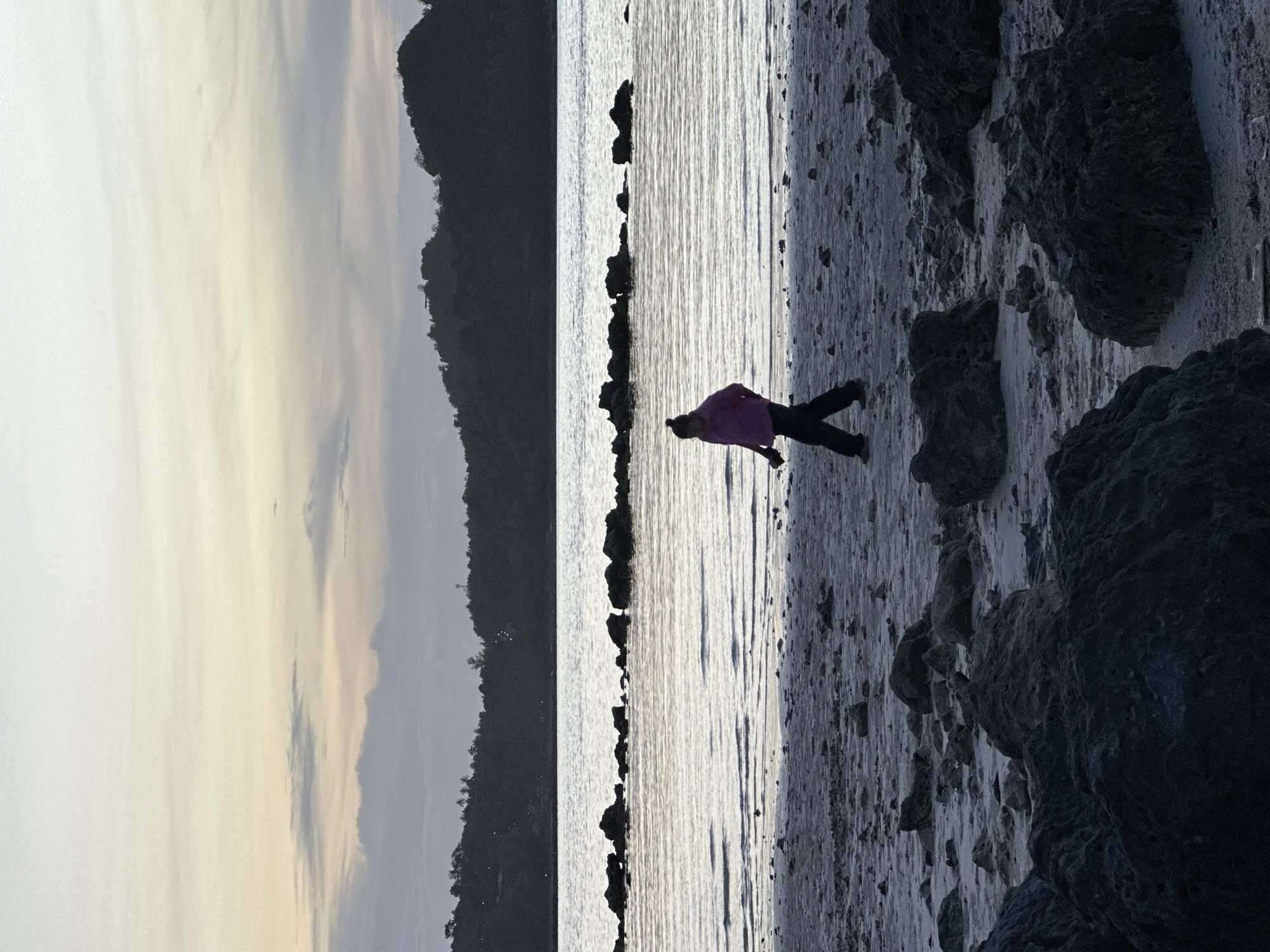 A lone figure, silhouetted in a pink jacket and dark pants, walks along a rocky, sandy beach at dusk. The figure is moving from left to right, facing away from the camera. The water is calm, with a line of dark rocks partially submerged in the middle ground. In the background, a dark, treed hillside rises, silhouetted against a pale sky with wispy clouds. The scene suggests a tranquil evening on the coast of Koh Phangan, Thailand, possibly in the Tambon Ko Phangan area.
