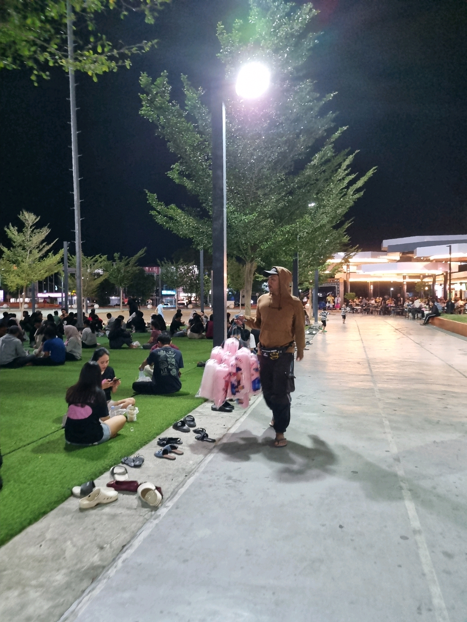 A group of people are gathered on a grassy area at night in Tanamodindi, Indonesia. A man in a brown hooded jacket and dark pants is walking on a concrete path. Several individuals are sitting on the grass, some of whom are looking at their phones. A few people are standing or walking in the background, near buildings with lights. There are trees and streetlights visible. Several pairs of shoes are scattered on the concrete path near the grassy area. The atmosphere appears to be relaxed and social, possibly a public park or gathering space.