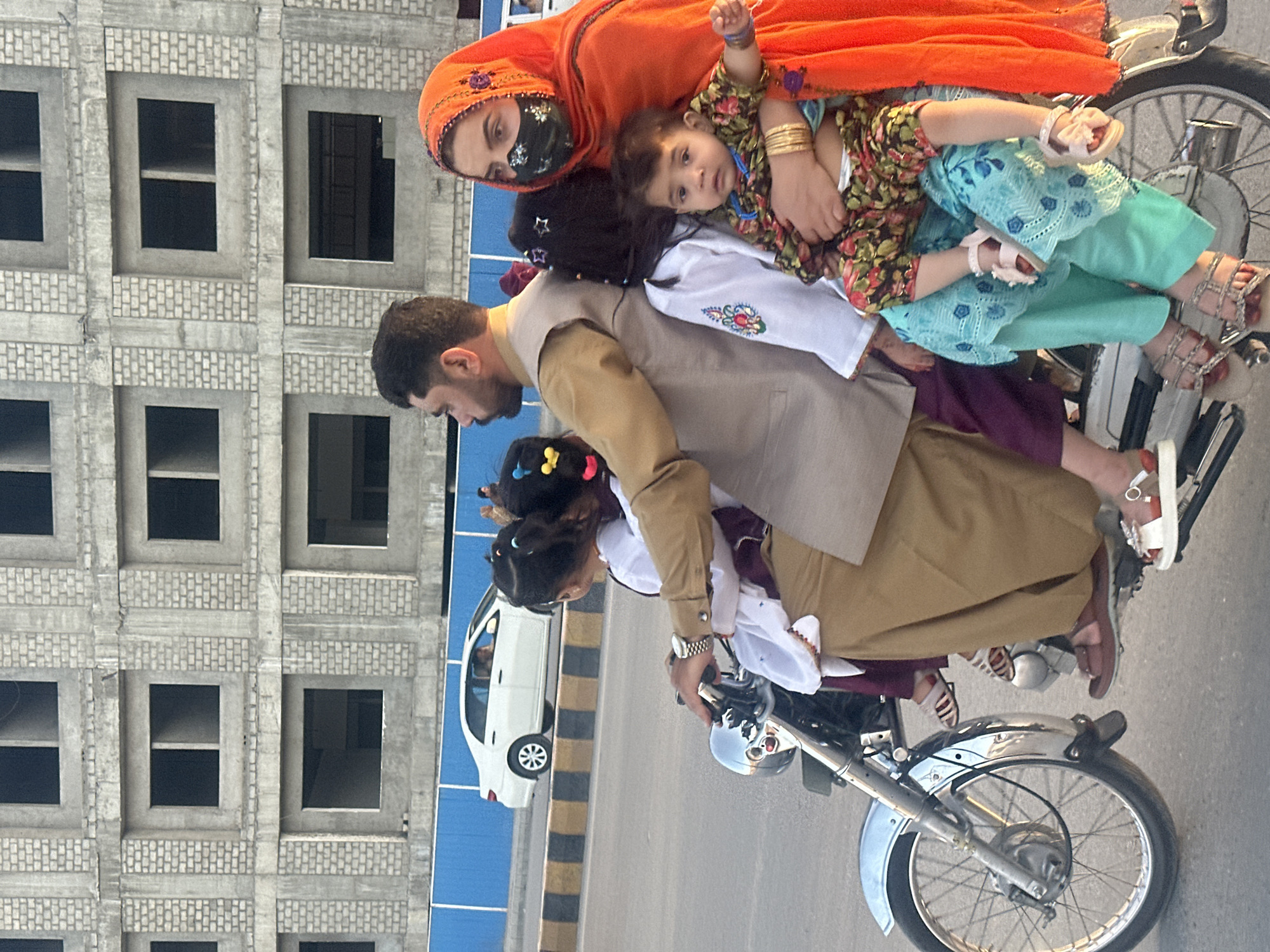 A family of five is seen riding a motorcycle on a road in Quetta City Tehsil, Pakistan. The father is driving the motorcycle, wearing a tan shirt and vest. His wife, wearing an orange headscarf and a black face mask, is sitting behind him and holding a baby dressed in a floral outfit and light blue pants and blouse. There are two more children riding between the parents. The road has blue and yellow safety barricades, and a white van is driving nearby. An unfinished grey concrete building is in the background.