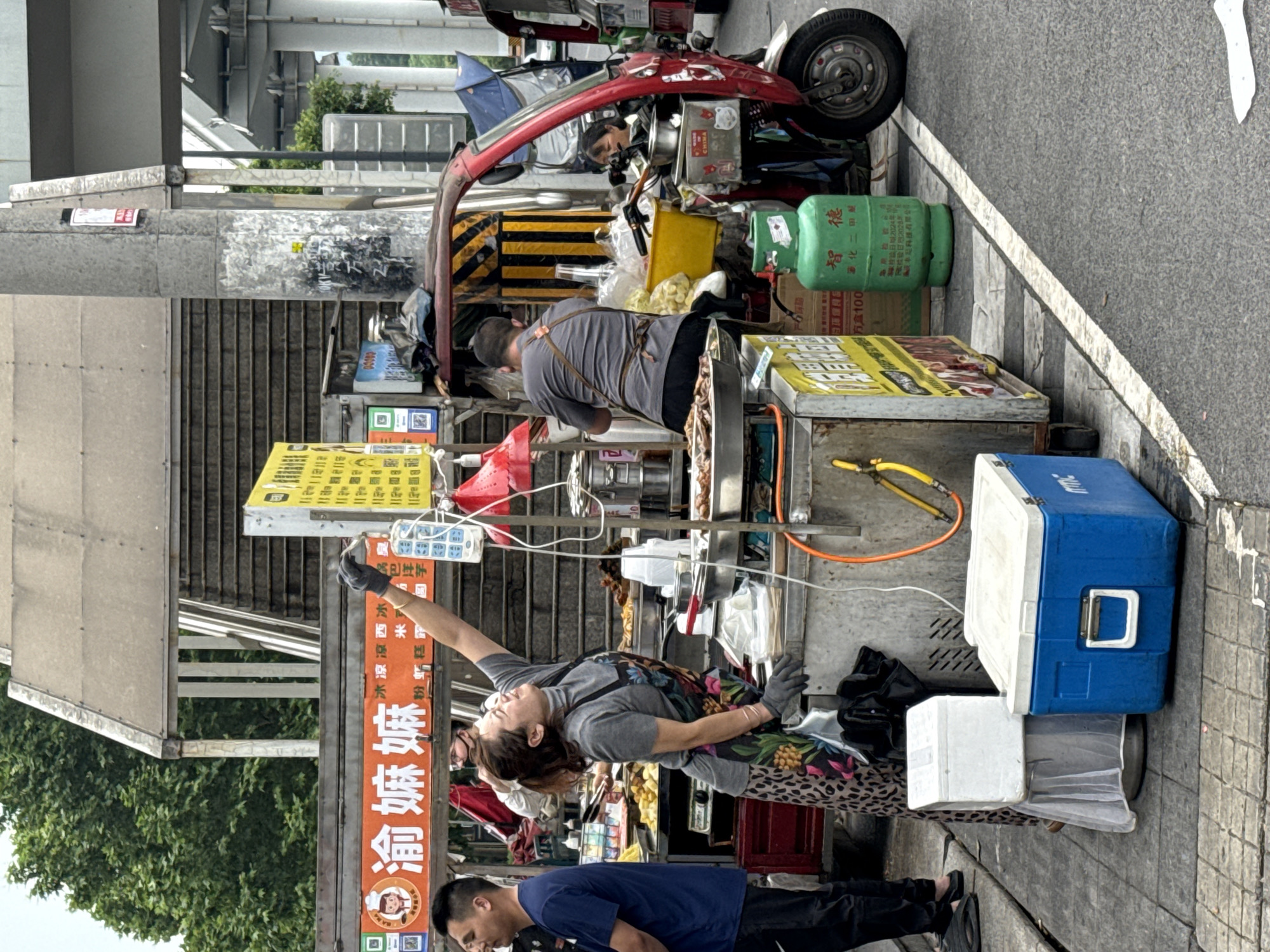 Here is a description of the image:
The image shows a street food vending setup in what appears to be the Chongqing High-tech Zone, China. A woman in a gray shirt and floral apron is reaching upward, presumably to adjust or grab something from above, while a man in a blue shirt stands in front of her, possibly a customer or associate. Another man is positioned at the back of the stall, seemingly preparing food.
The vending setup involves a variety of objects and equipment, including cooking equipment with food skewers, a blue and white cooler, various containers, and a red three-wheeled vehicle cluttered with goods. A green gas cylinder is also visible.
A prominent sign with Chinese characters "●渝嫲嫲" is visible, along with some food descriptions. The environment appears to be a public walkway with a street adjacent to it. There are trees and a building structure visible in the background. The scene suggests a bustling street food market or vendor area.